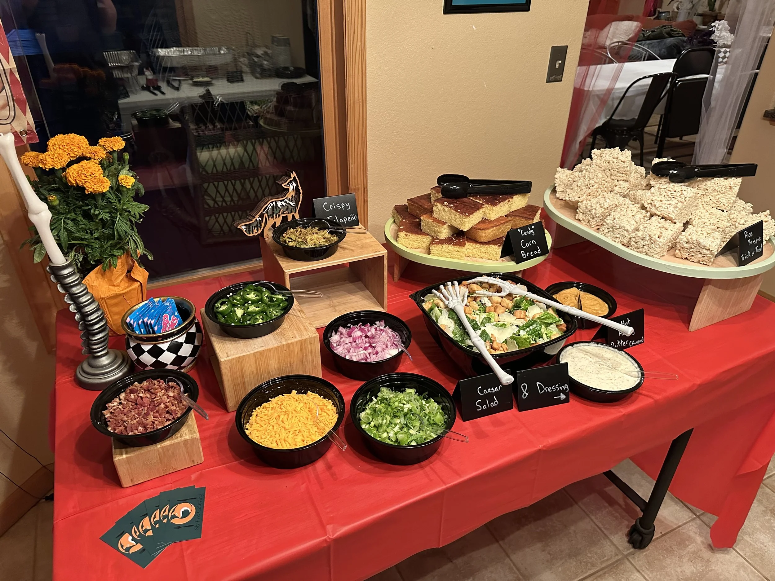 A buffet table with various salads and side dishes, including chopped onions, sliced jalapeños, shredded cheese, and Caesar salad, along with bread and rice Krispies treats, decorated with a pumpkin, a candle, and small decorative signs.