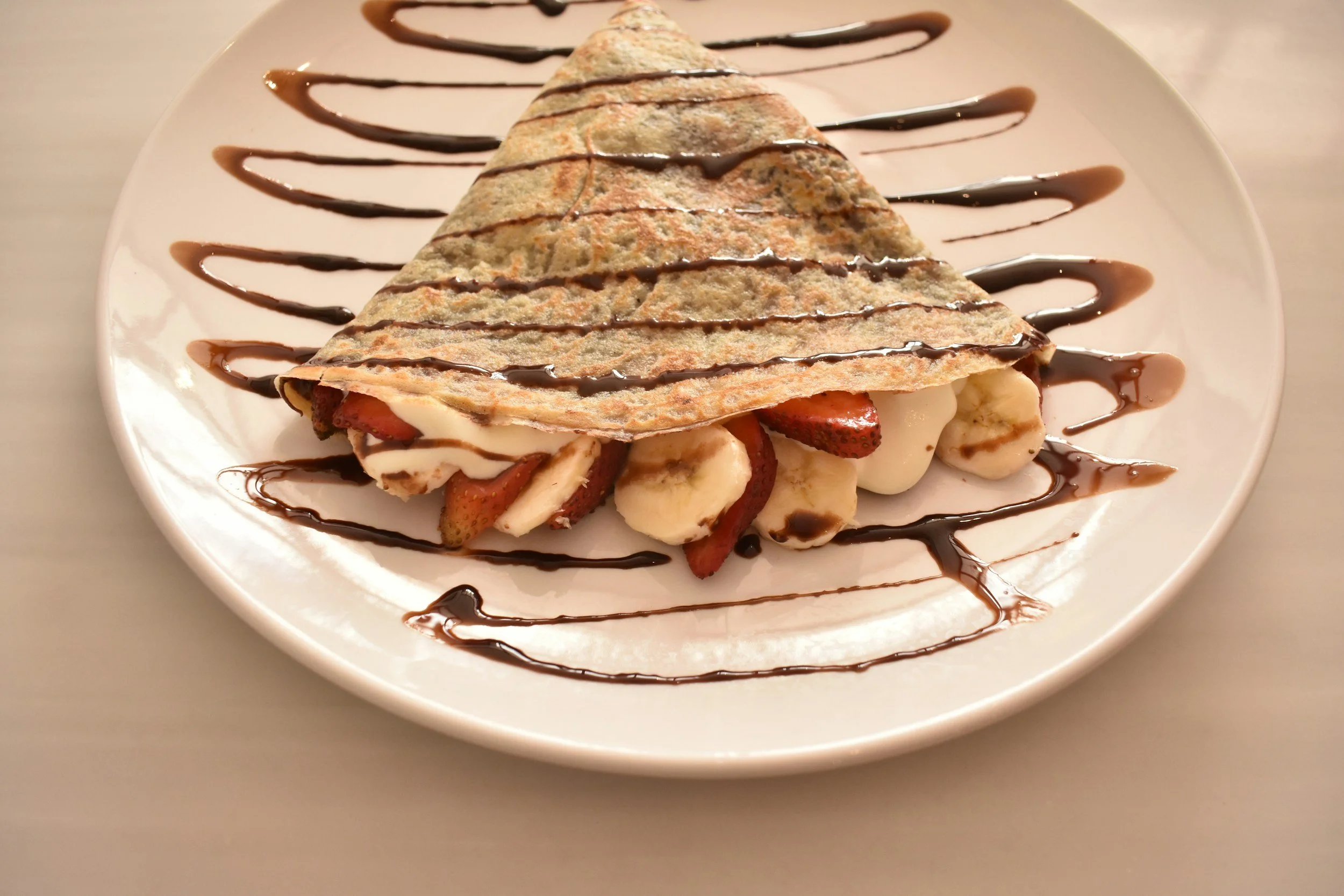 A crepe filled with sliced strawberries, banana slices, and whipped cream, drizzled with chocolate sauce.