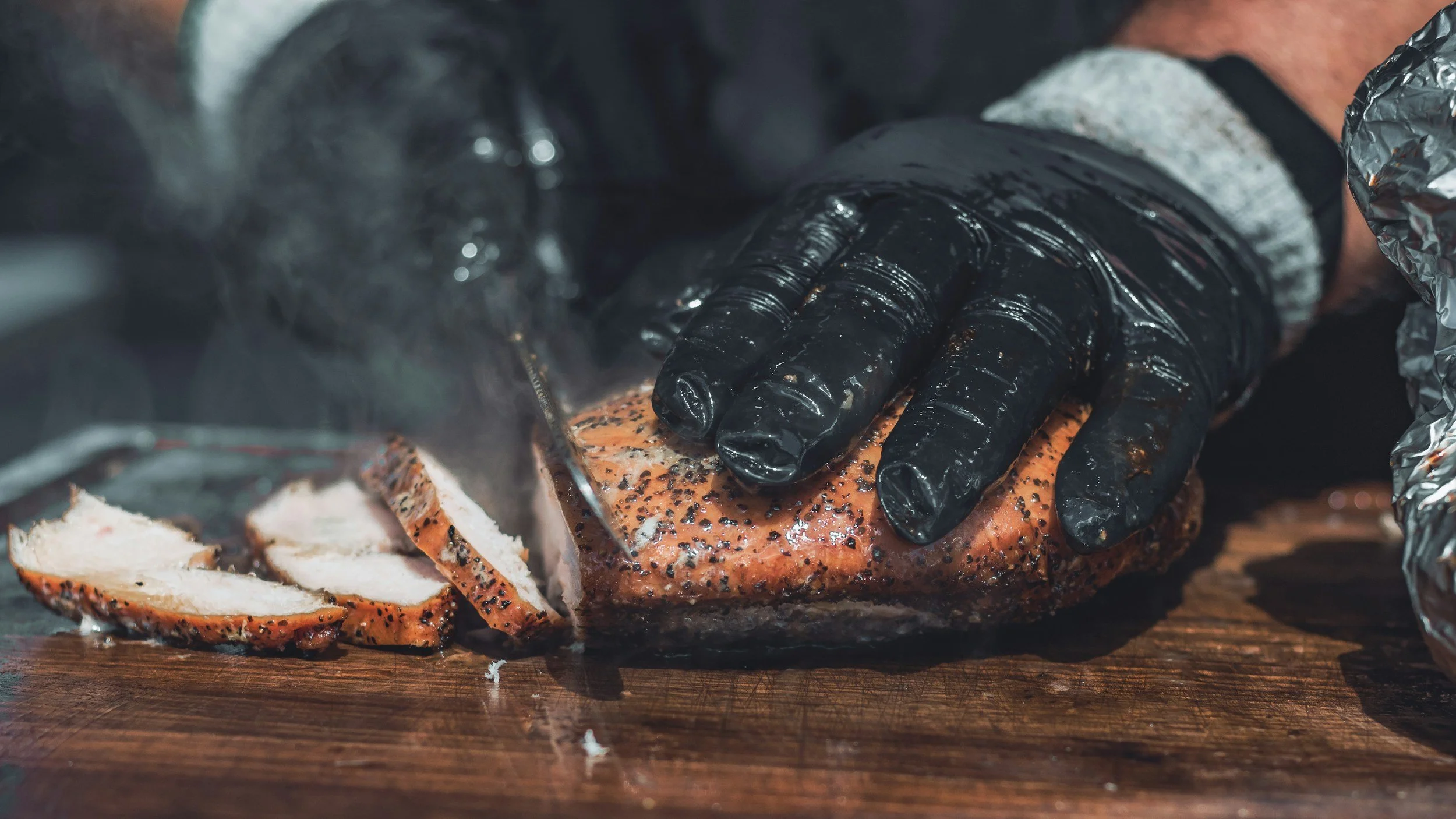 A person using a gloved hand slices smoked salmon on a wooden cutting board.