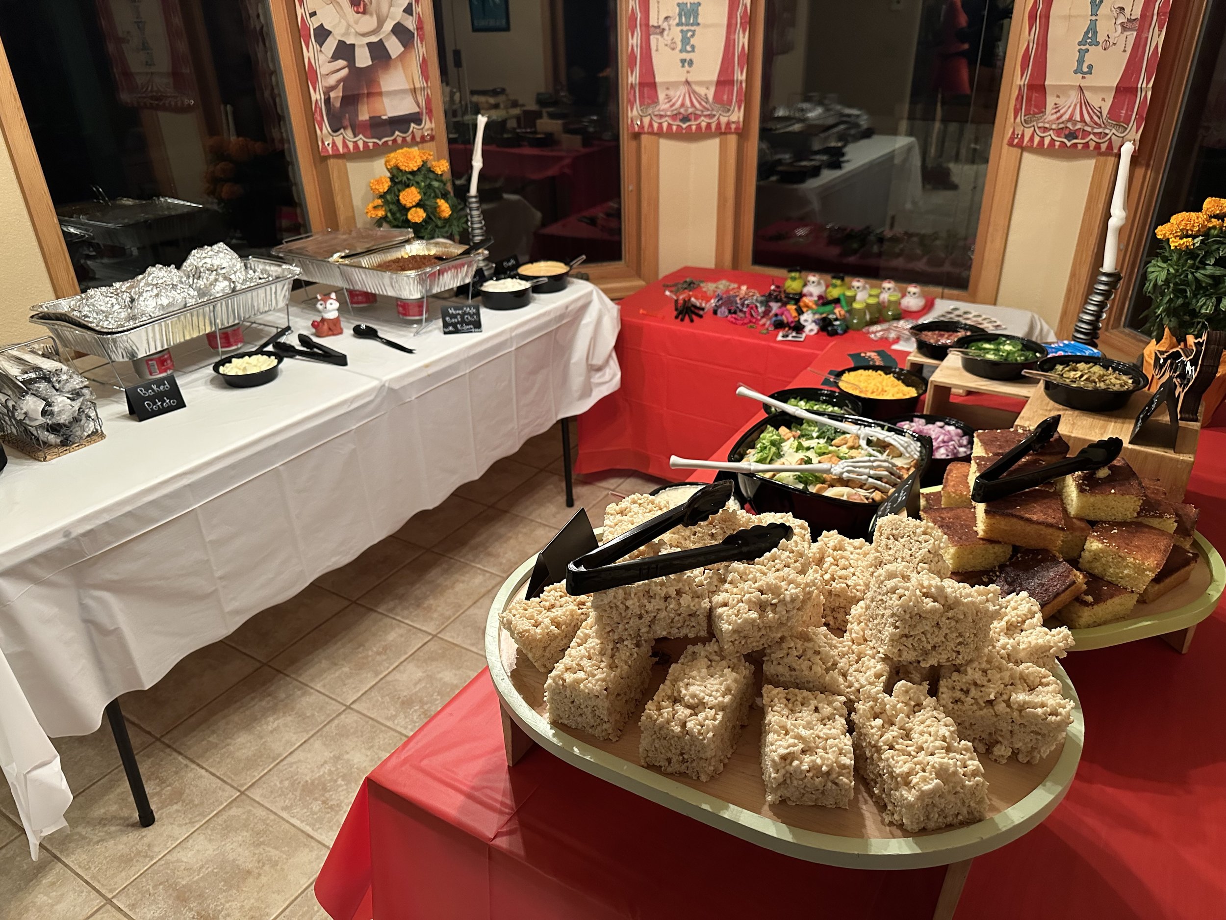 Buffet table with various dishes including rice crispy treats, salad, baked potatoes, and other dishes, decorated with Halloween themed decorations and toys.