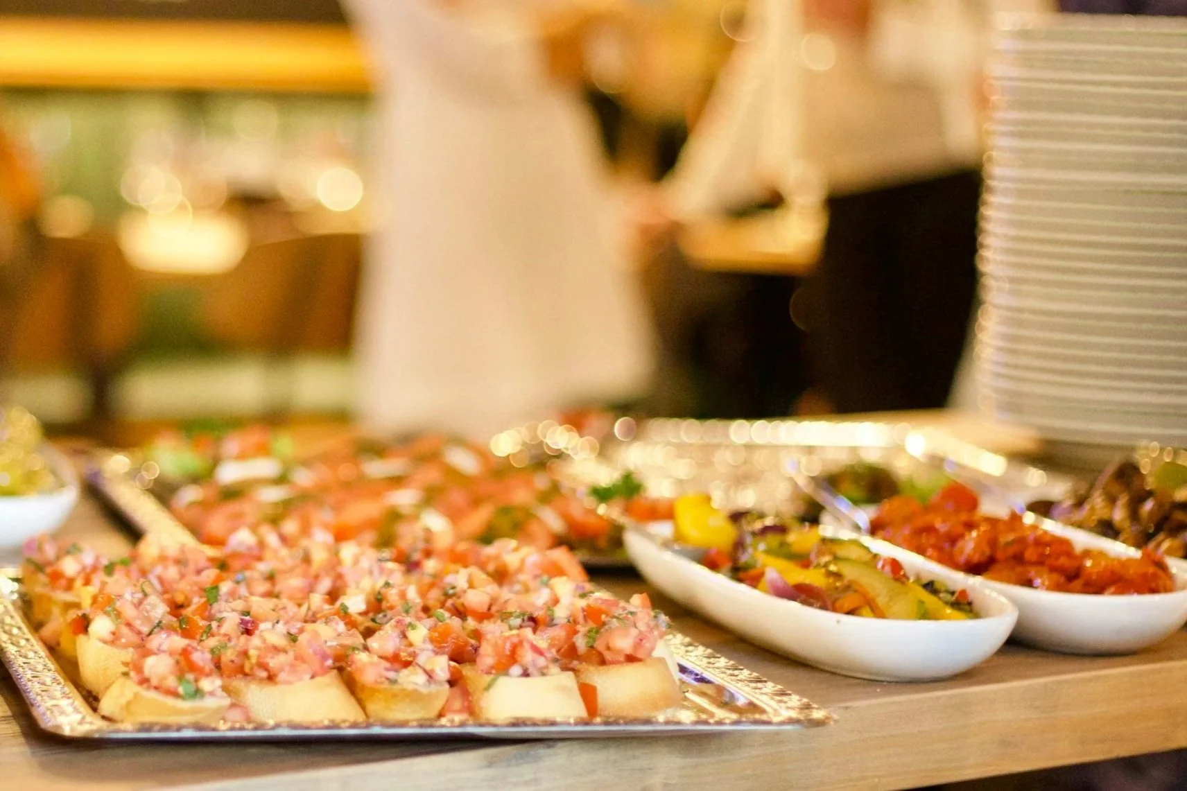 A buffet table with various dishes, including a tray of nachos with chopped tomatoes, and bowls of grilled vegetables and saucy chicken.