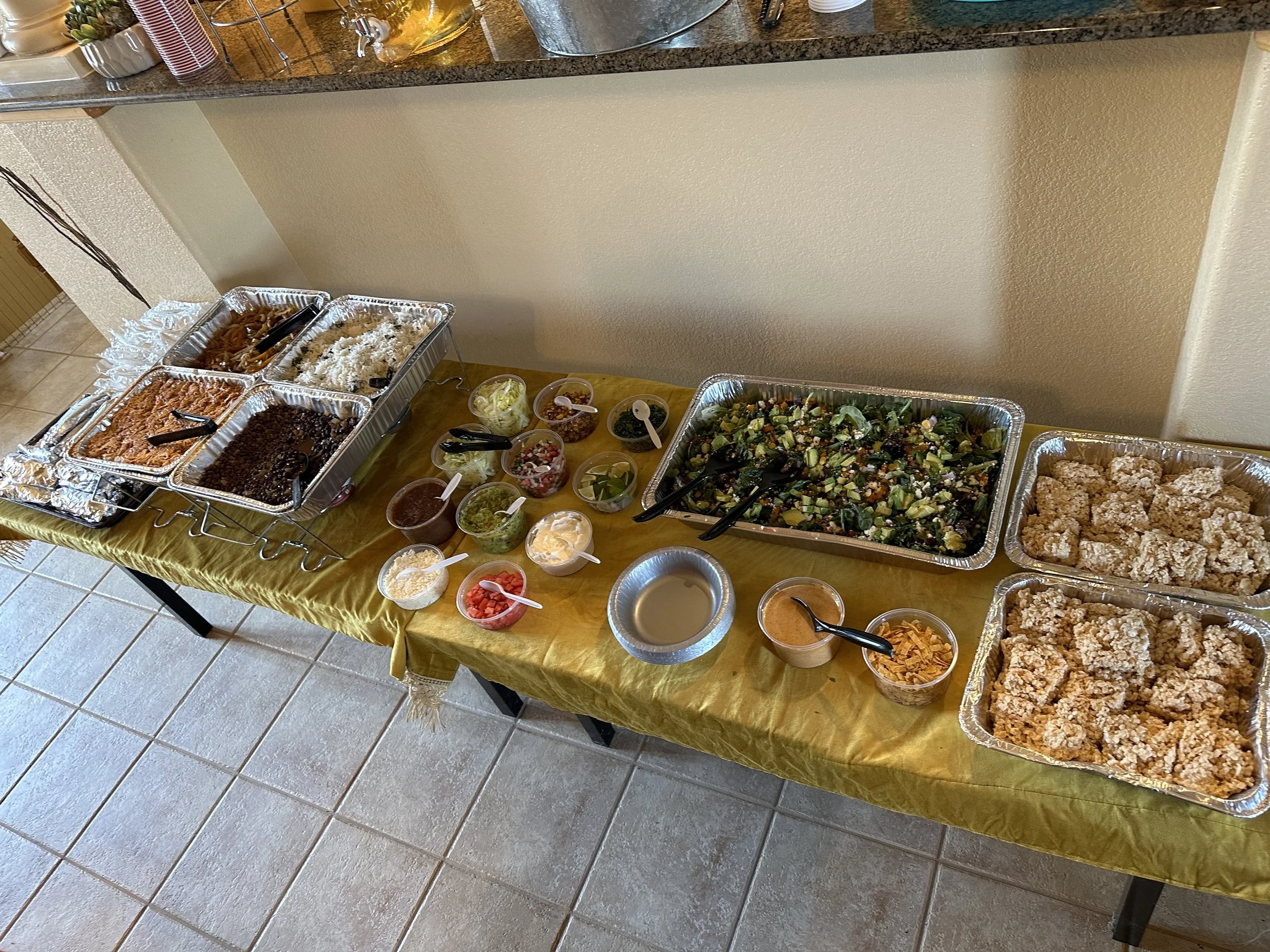 Buffet table with various foods including salads, fried chicken, rice, beans, sauces, and toppings in small bowls, covered with aluminum trays and containers, set on a yellow tablecloth.