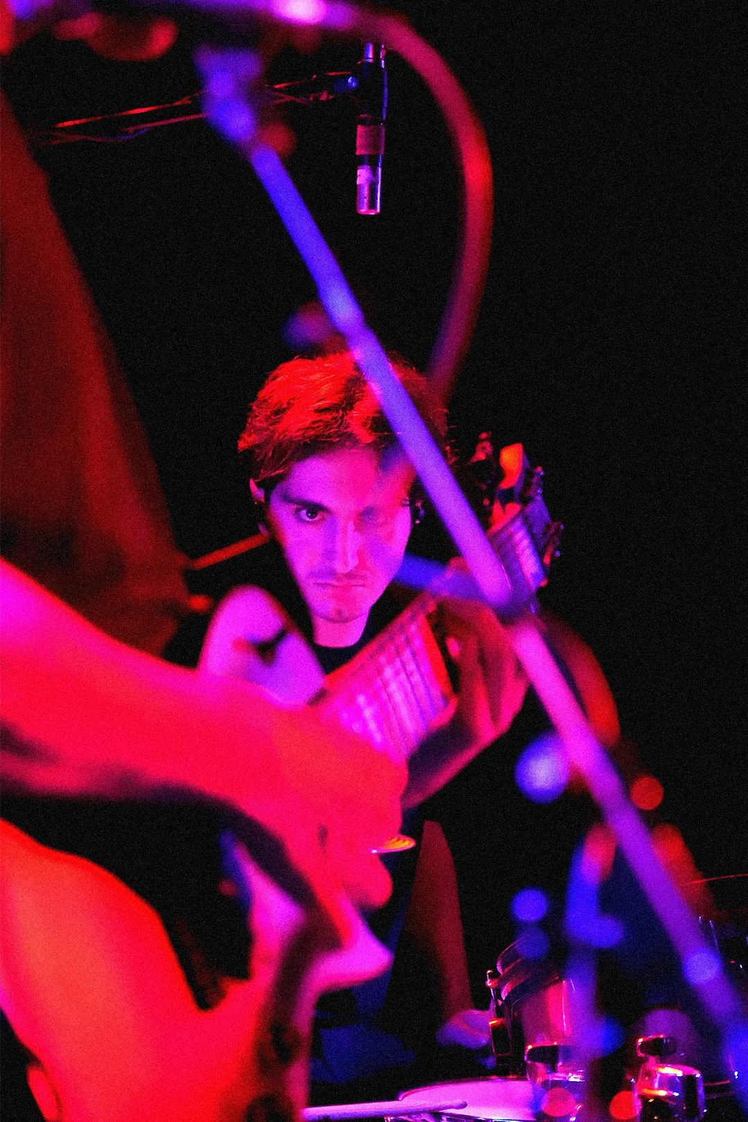 A man with red hair playing an acoustic guitar on stage, illuminated by purple and pink stage lights, with a microphone overhead and various musical equipment visible.