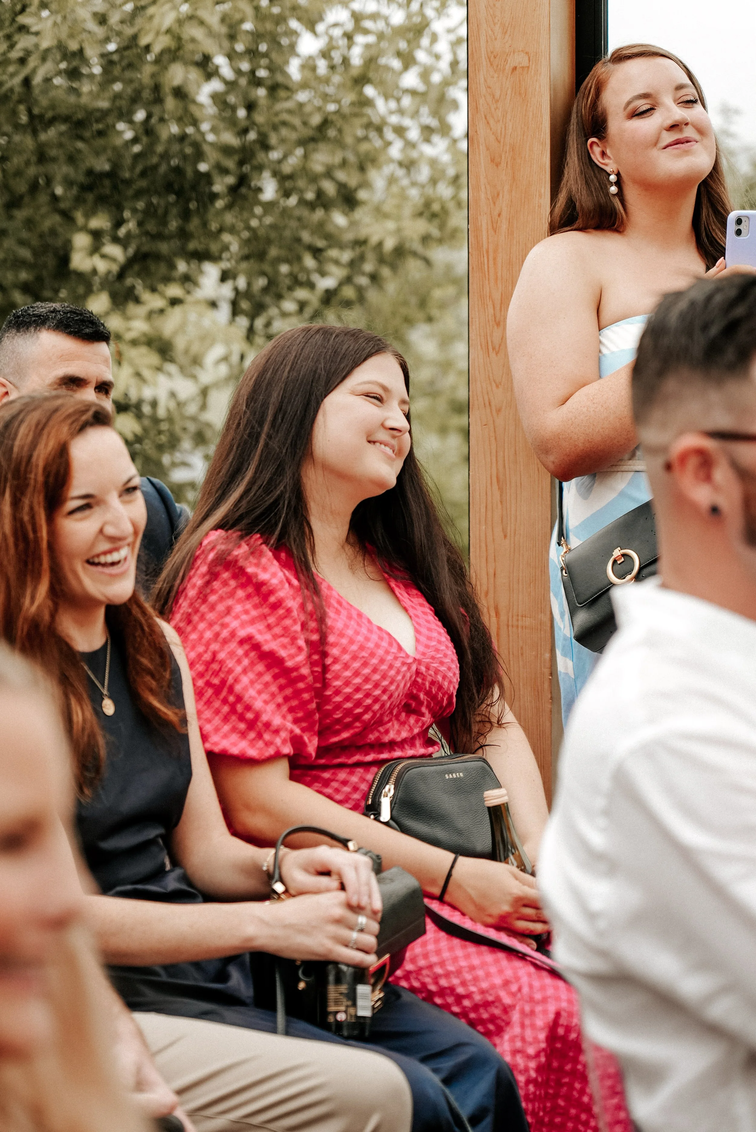 Group of people at an outdoor event, some smiling and enjoying the moment, one woman in a red dress seated in front, others around her, some standing and some sitting, with a background of trees.