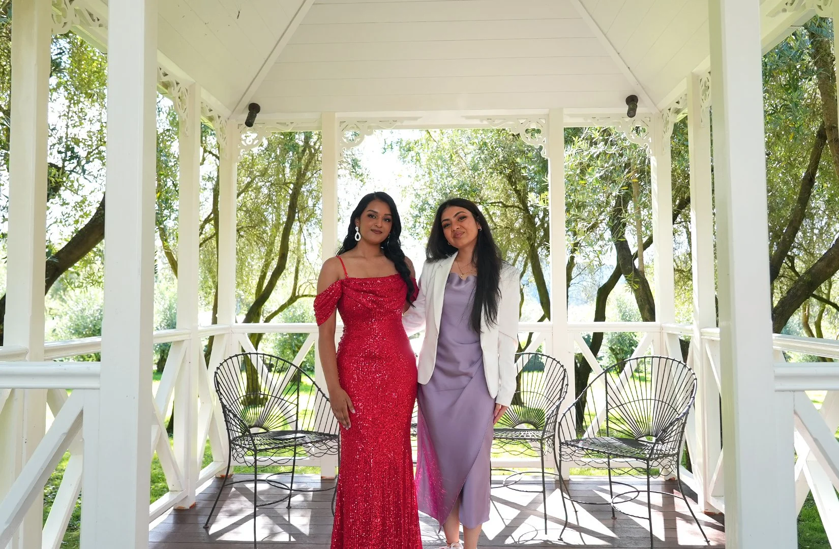 Two women in formal attire standing on an outdoor white gazebo with trees in the background.