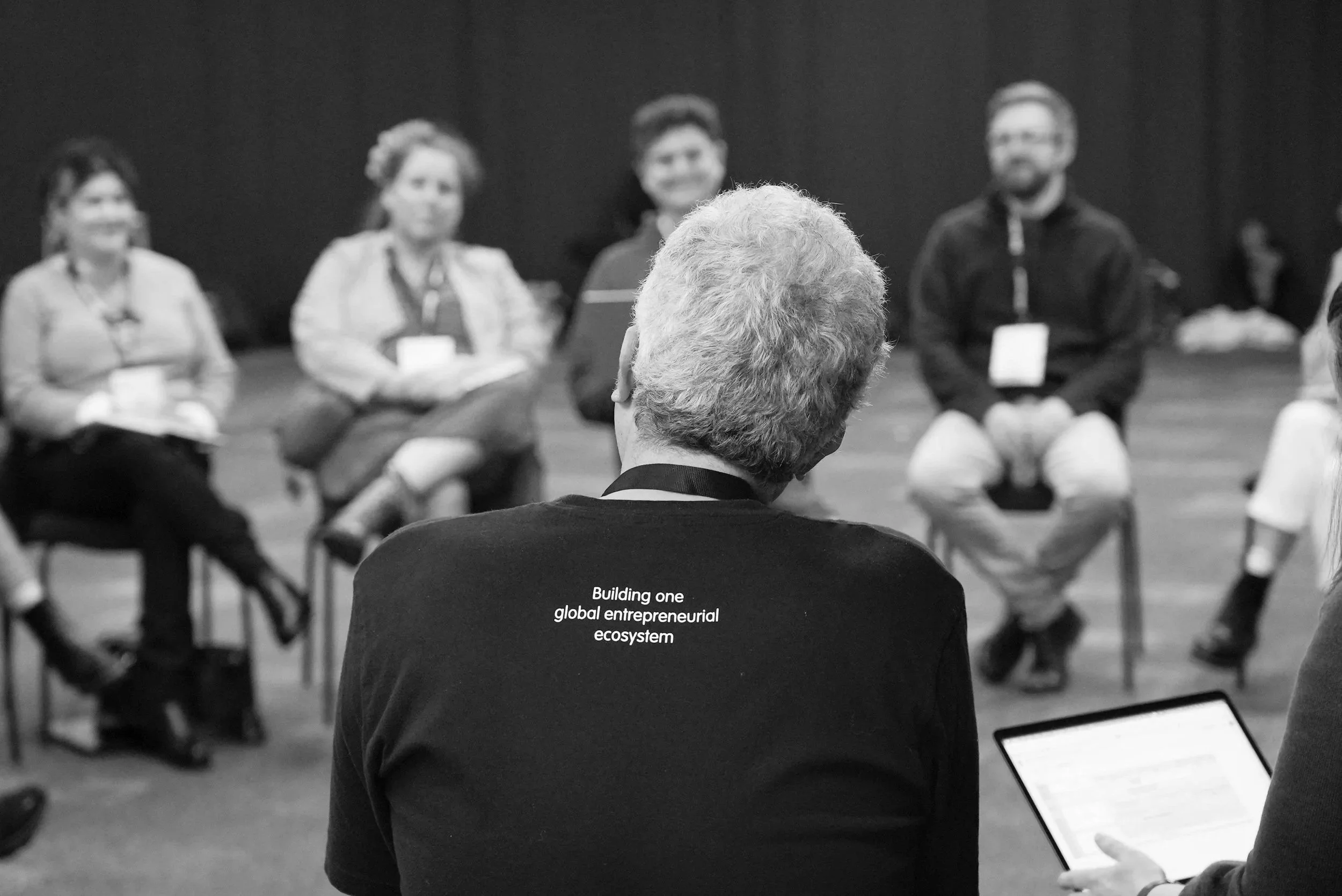 An older man with gray hair seen from behind, wearing a black shirt with the words 'Building one global entrepreneurial ecosystem' printed on the back, is attending a panel discussion with five people seated in front of him. The scene is in black and white.
