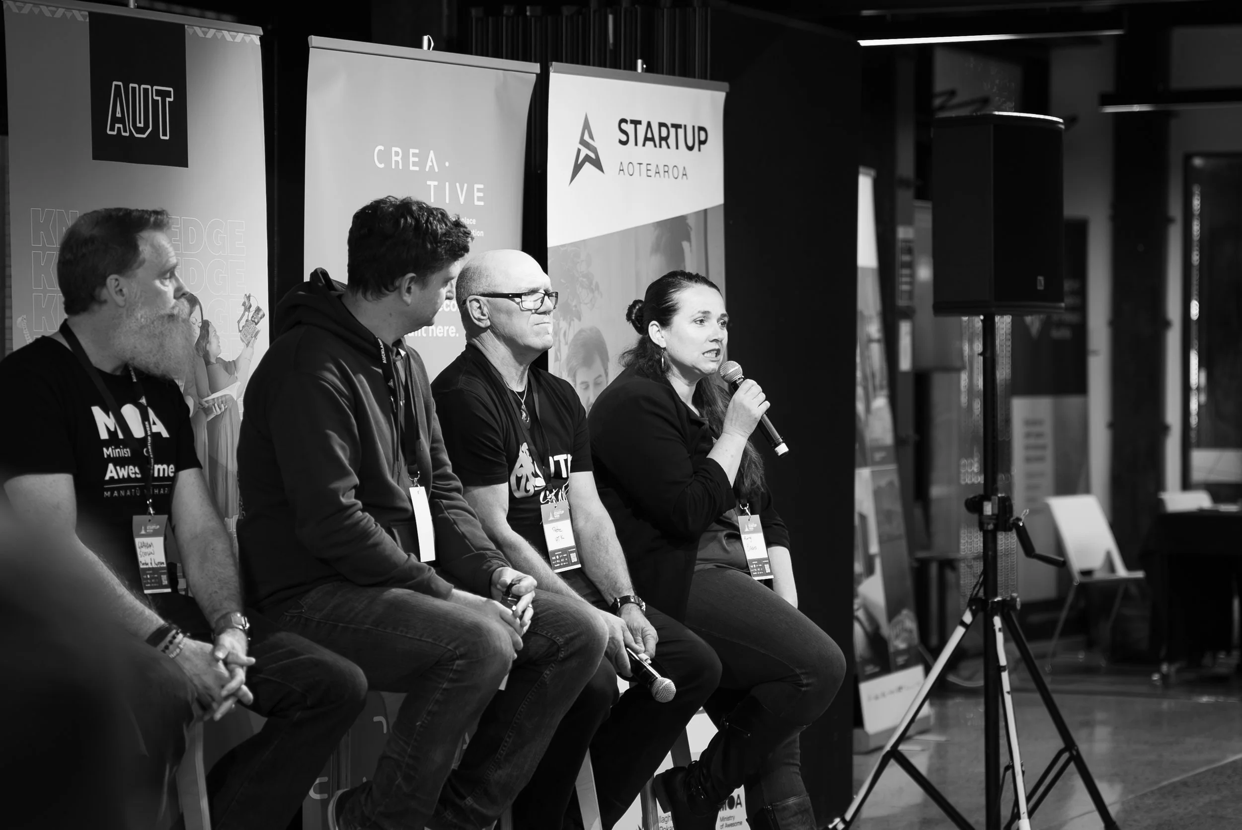 A panel discussion at a startup event, with four people seated on a stage, one woman speaking into a microphone, and banners in the background with startup-related logos and text.