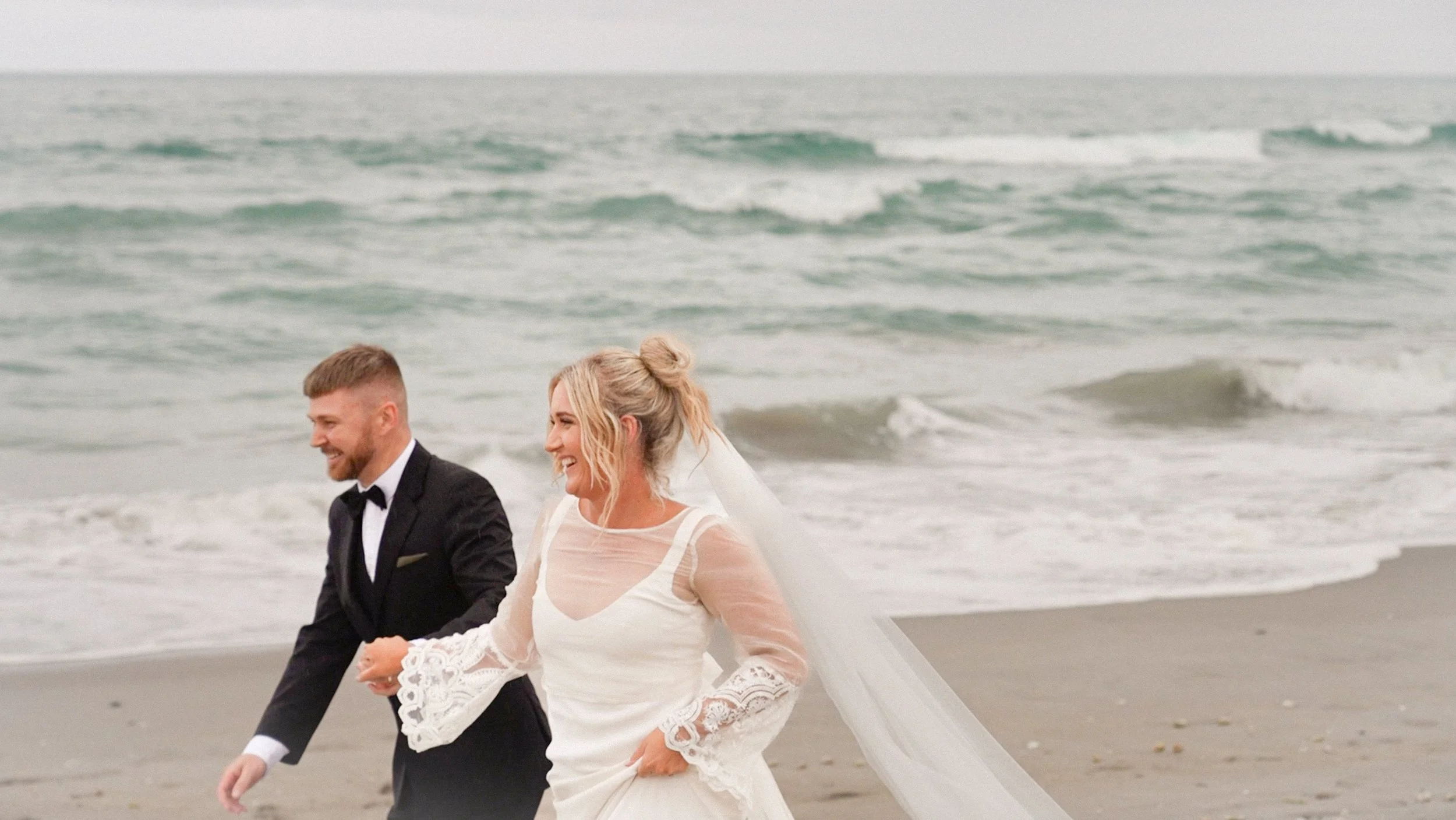 A happy bride and groom walking along the beach, smiling and holding hands, with ocean waves in the background.