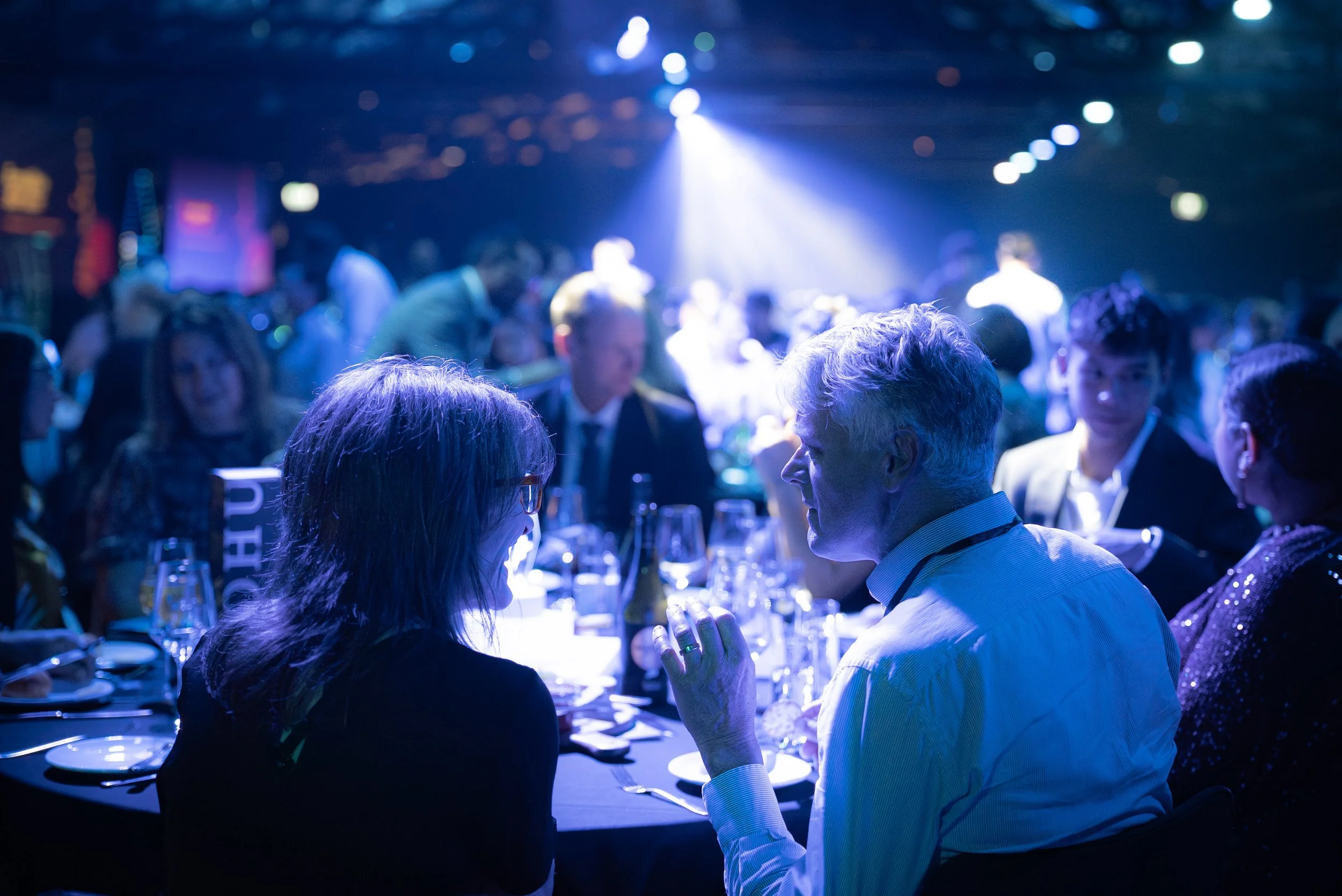 People seated around a table at a formal event or party, illuminated by blue lighting with a bright spotlight in the background.