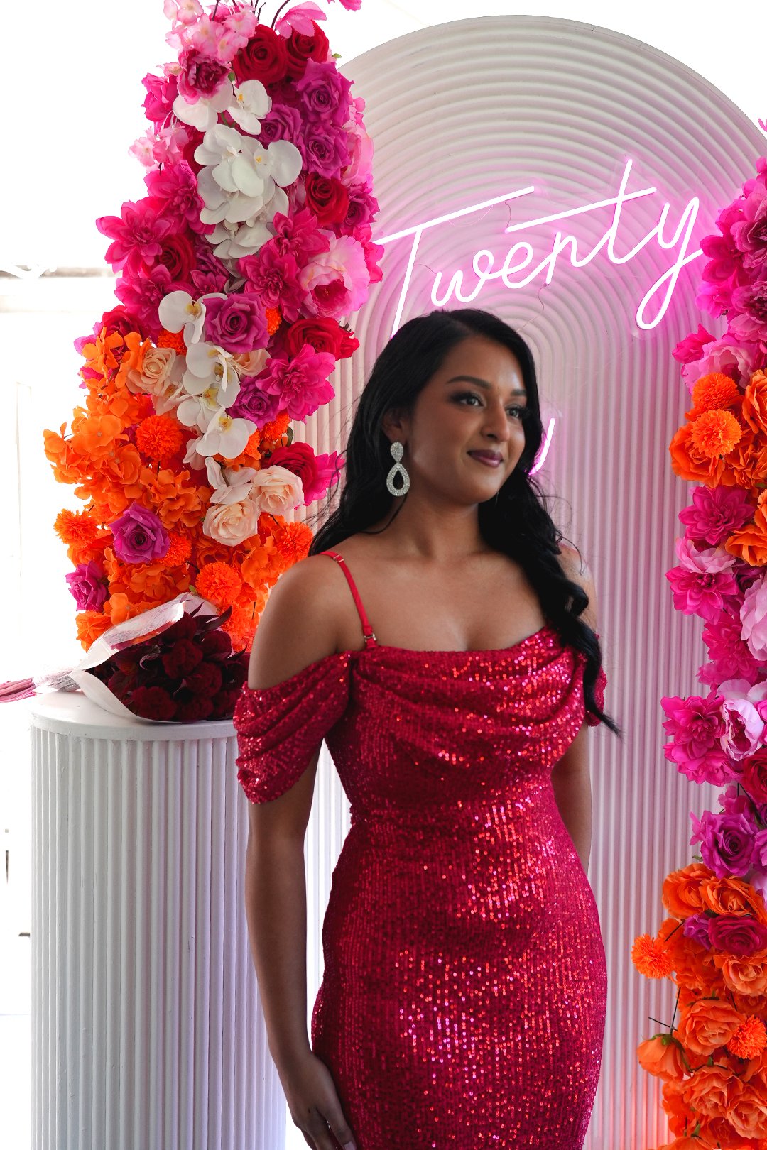 A woman in a red, sequined dress standing in front of a floral backdrop with pink and orange flowers and a neon sign that says "Twenty".