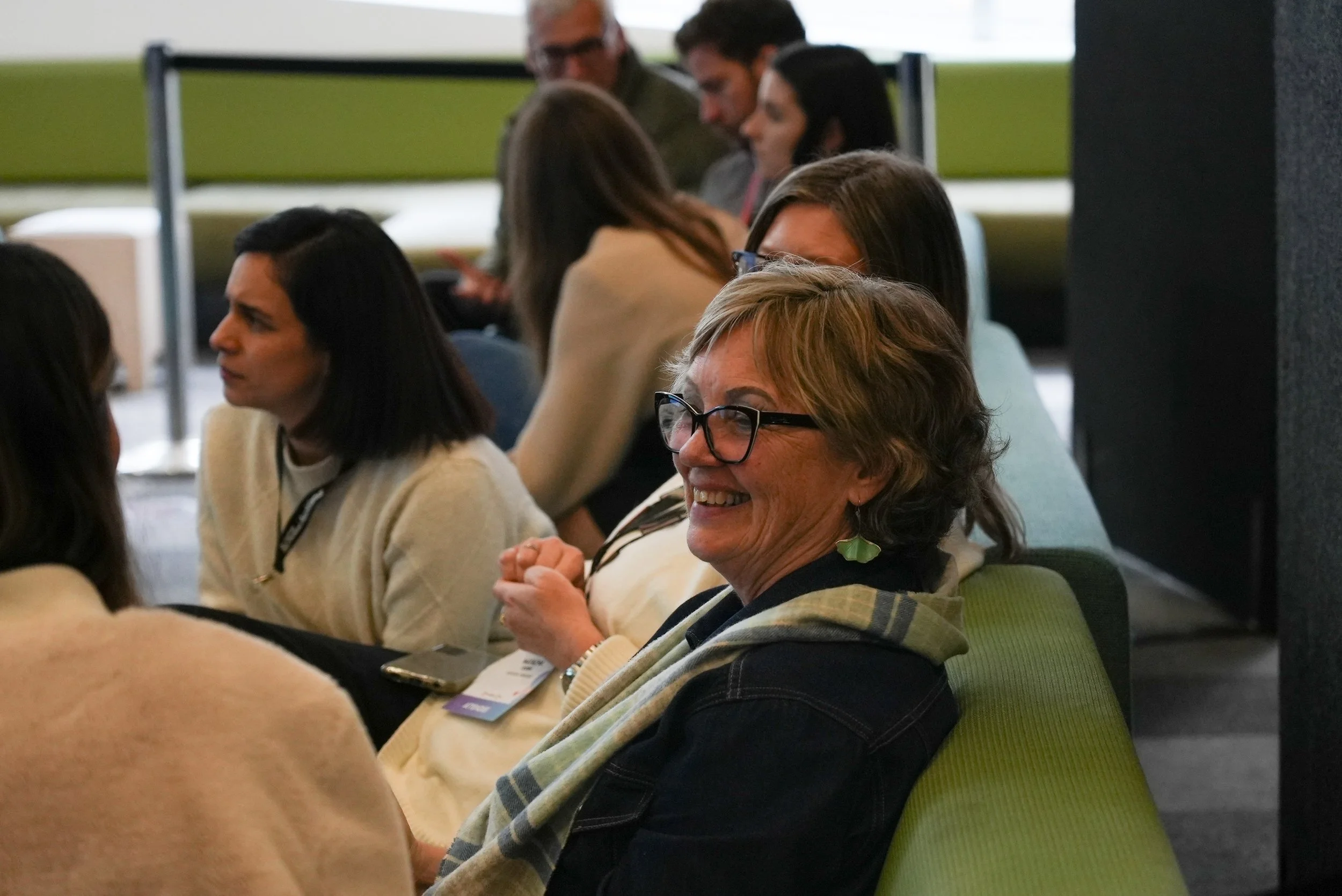 A group of diverse people sitting in a conference room, engaging in conversation. The focus is on a smiling older woman with glasses and earrings, seated on a green chair at the forefront.