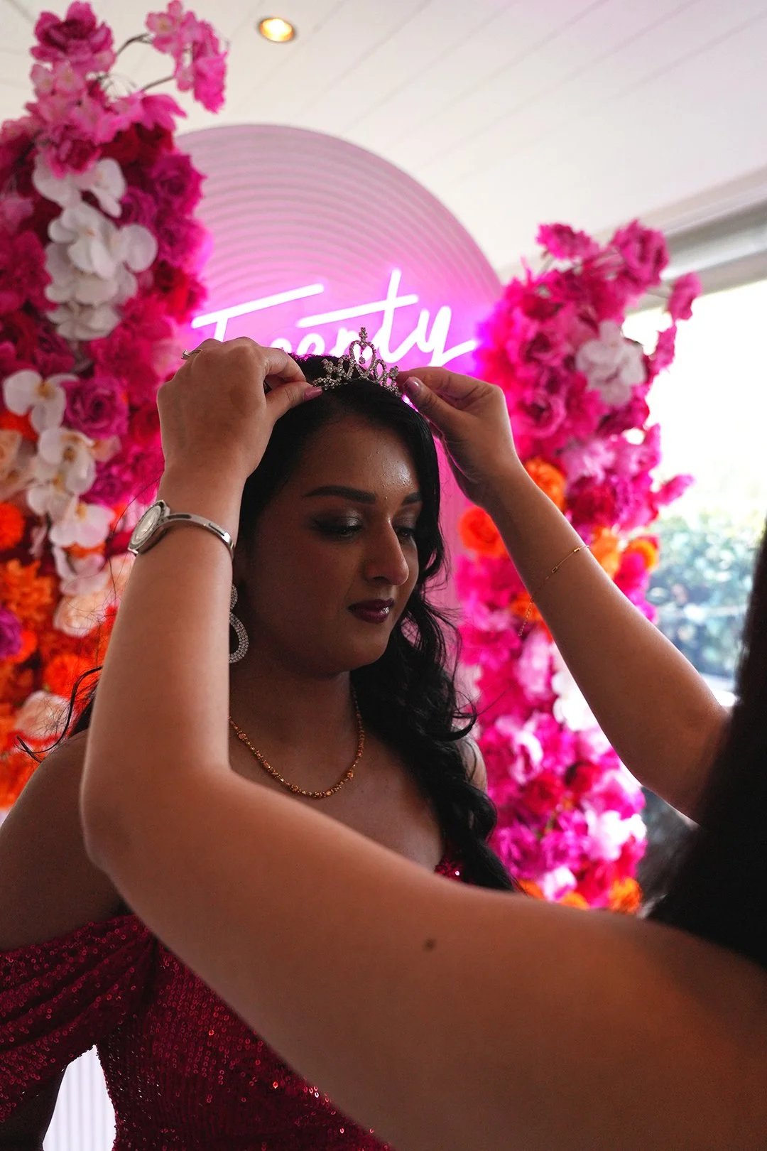 A woman is being crowned with a tiara at a celebration, standing in front of a pink floral backdrop and a neon sign that says "Beauty."