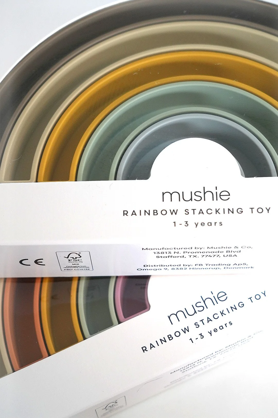 Close-up of packaging for Mushie rainbow stacking toy designed for children aged 1-3, featuring colorful concentric rings visible through the clear window of the box.