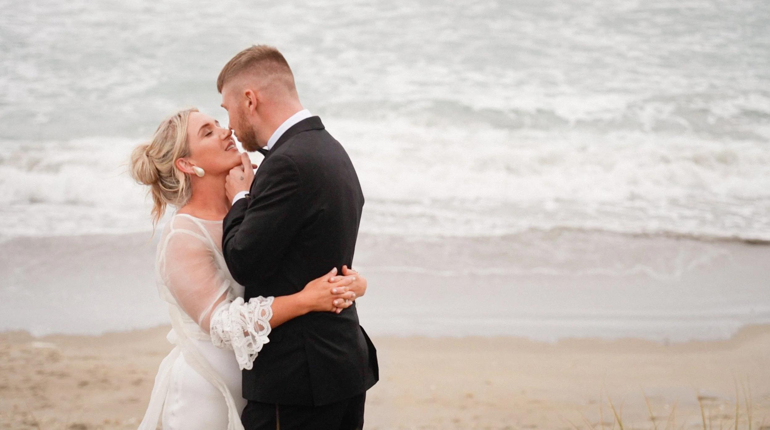 A couple in wedding attire embracing on a beach, with the ocean and sand in the background.