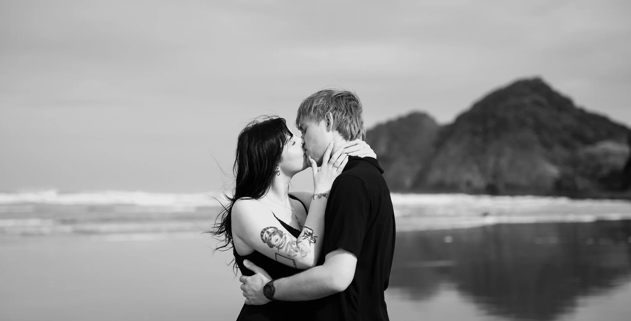A black and white photo of a couple kissing on the beach, with a rocky hill in the background.