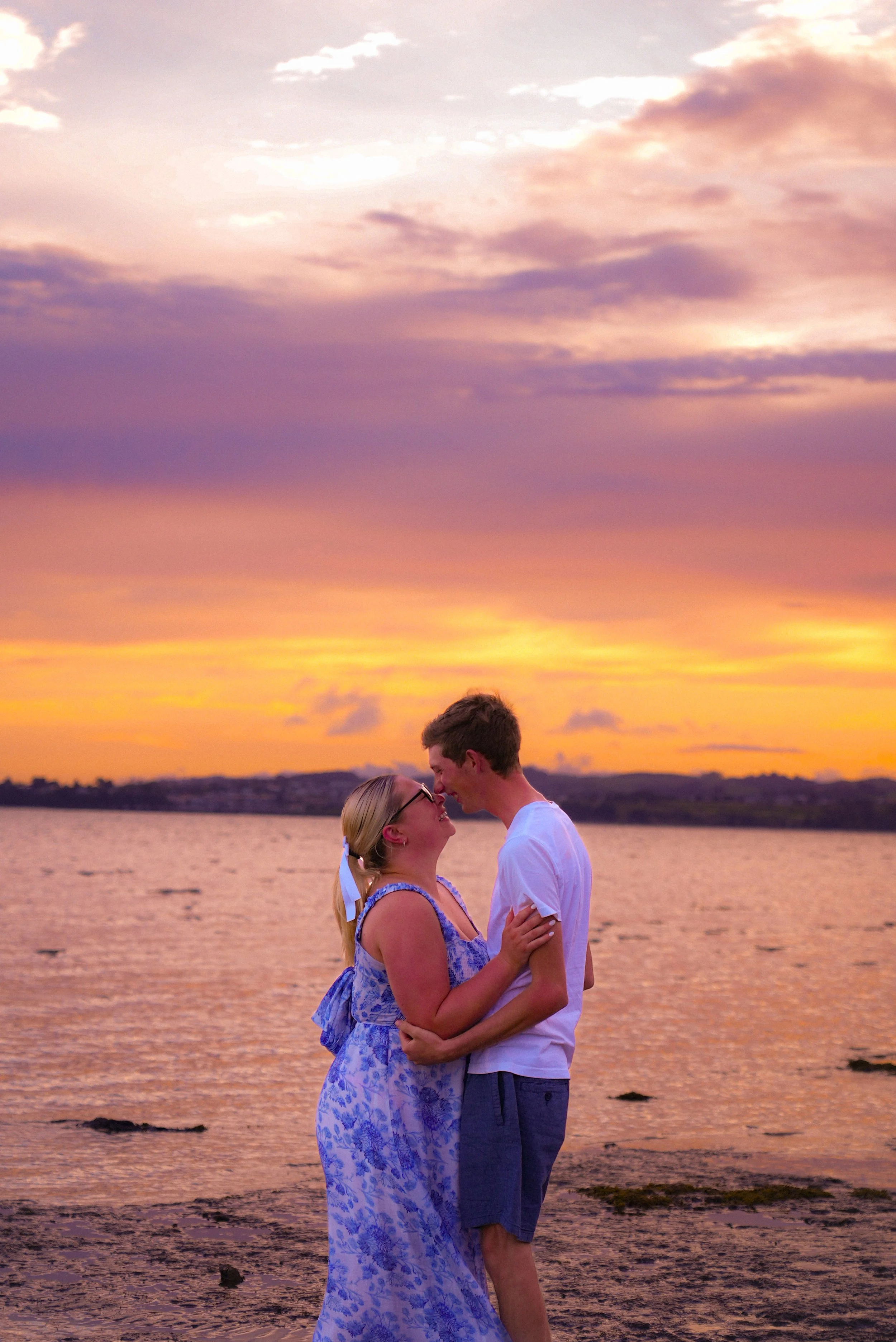 A couple embracing by the water at sunset, with a colorful sky and calm water in the background.