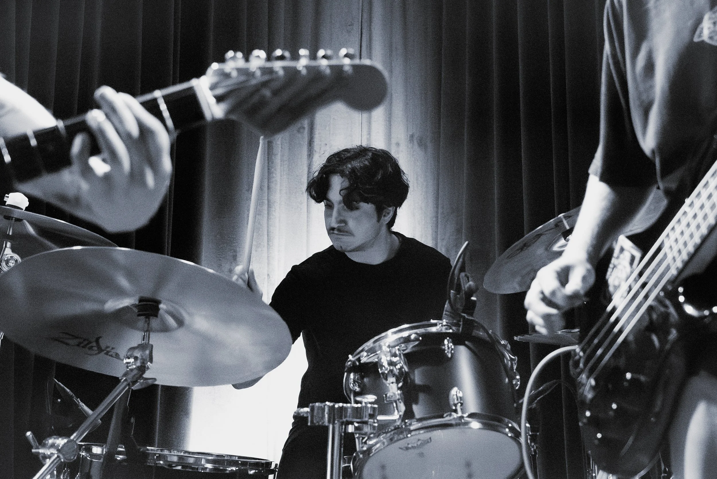 A black and white photo of a young man with dark curly hair playing a drum set in a room with dark curtains. Parts of other musicians playing guitar and bass are visible.