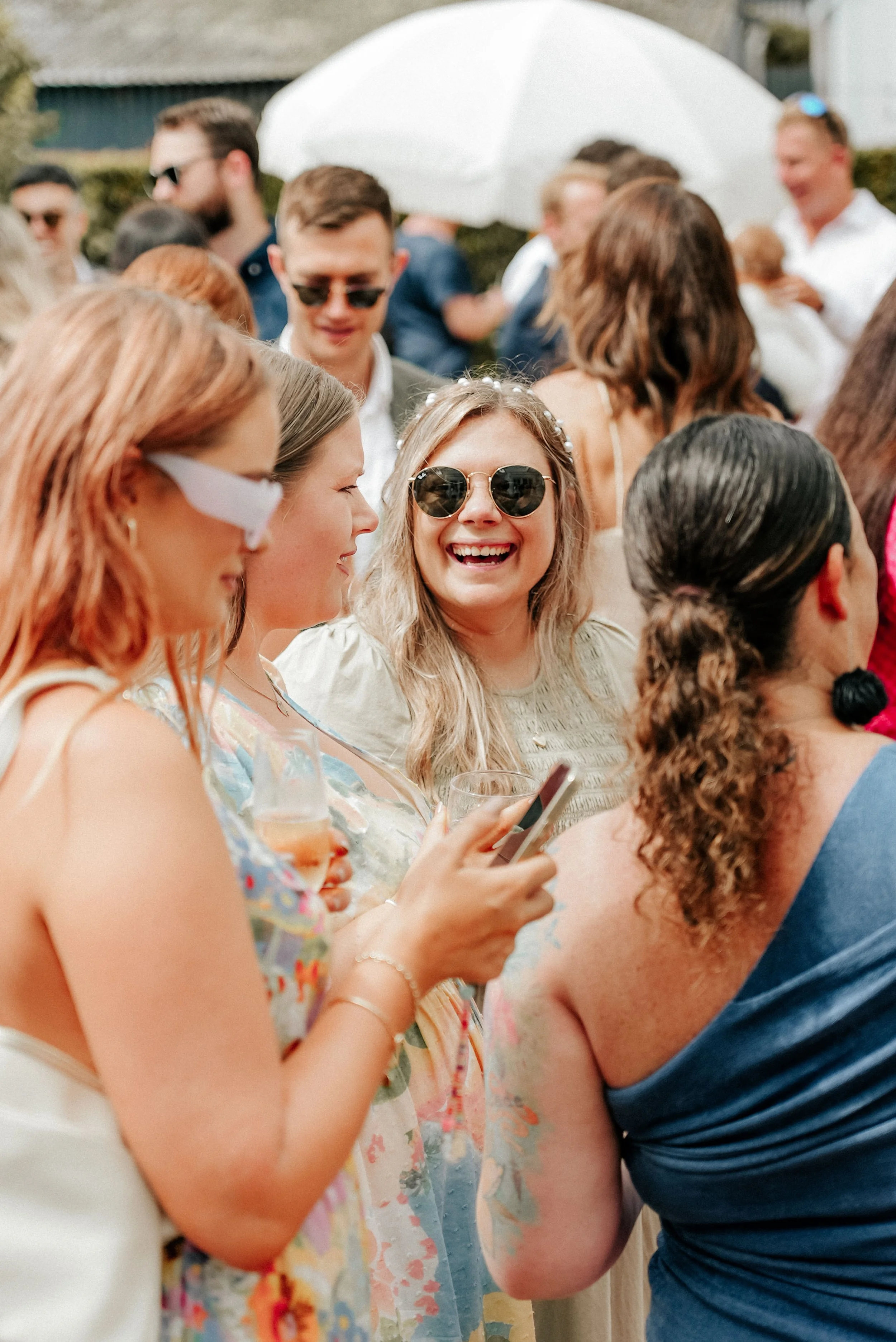 Group of people at an outdoor gathering, some wearing sunglasses, with one woman in the center smiling and holding a drink and phone.