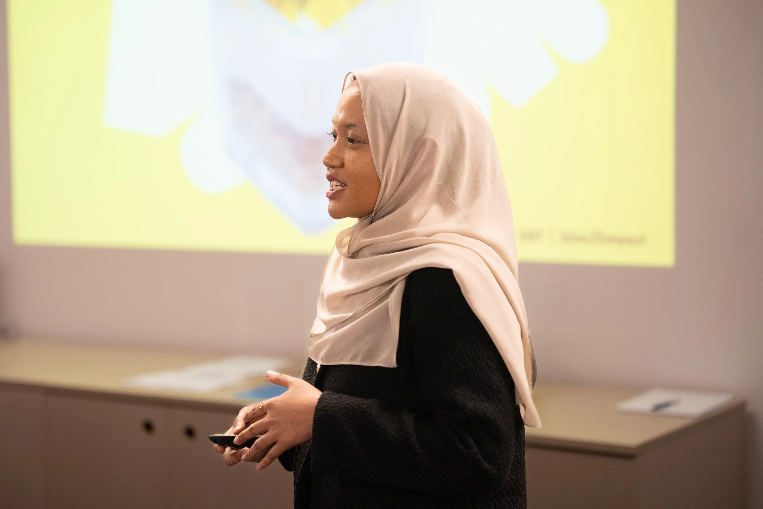 A woman wearing a beige hijab and black outfit giving a presentation in front of a yellow slide.
