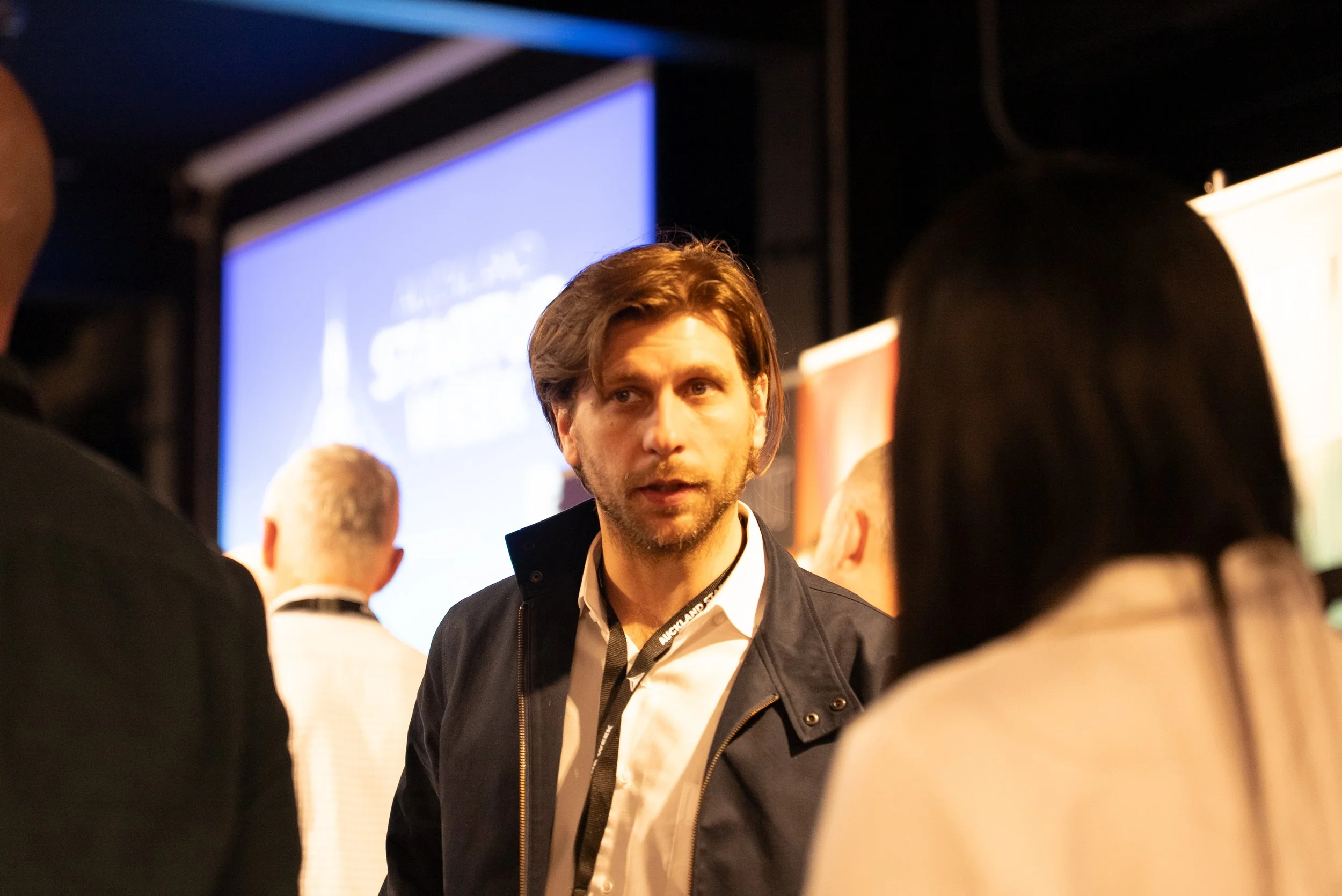 A man with brown hair and a beard engaged in conversation at a conference or event, with a group of people around him and a blurred blue presentation screen in the background.
