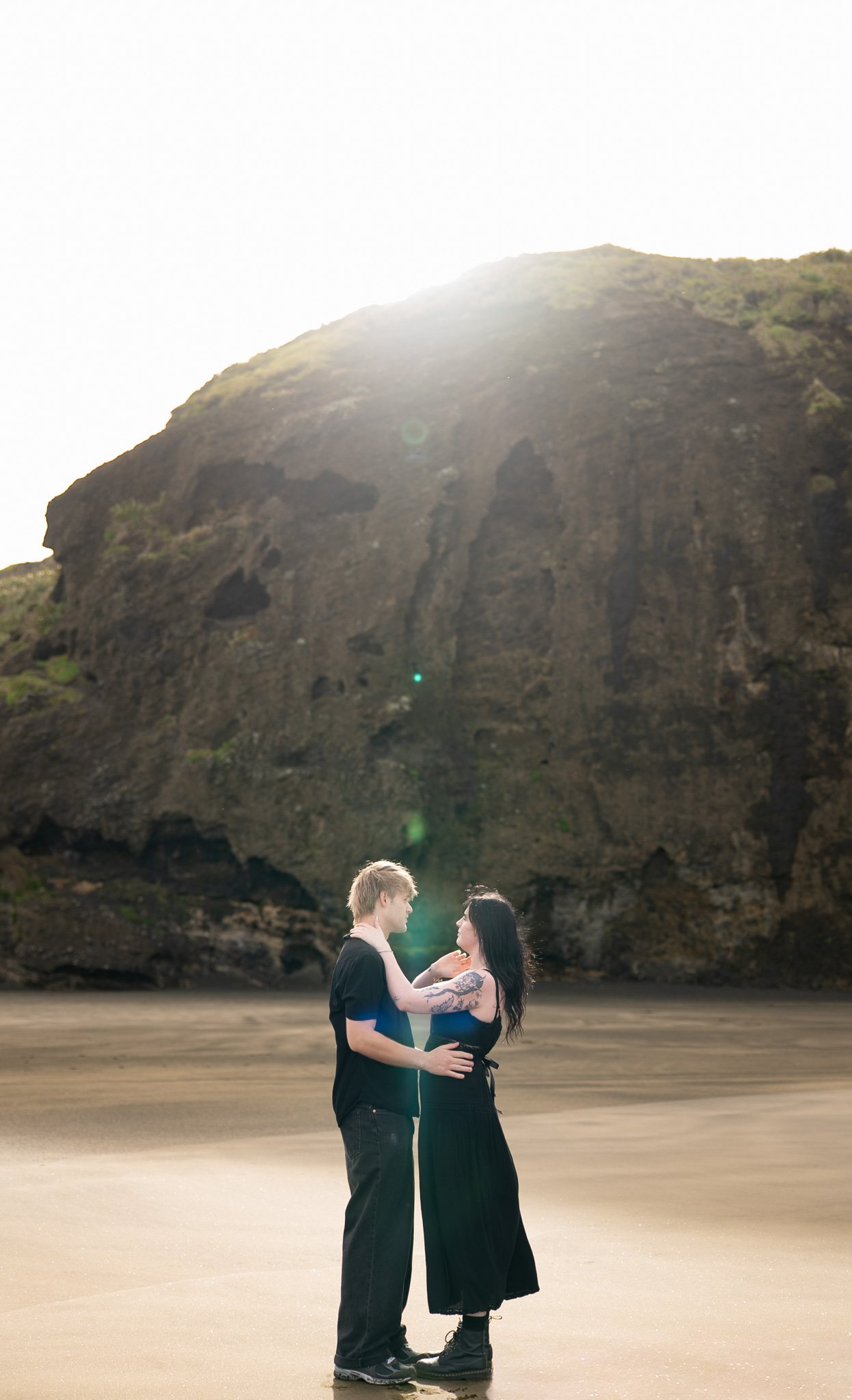 A couple standing on a beach, embracing each other, with a rocky cliff in the background and bright sunlight overhead.