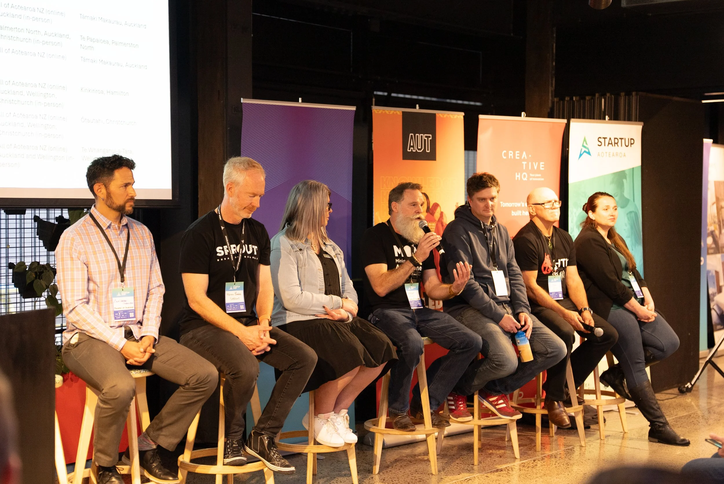 A panel of seven people sitting on stools on a stage, with colorful banners behind them, at a conference or event.