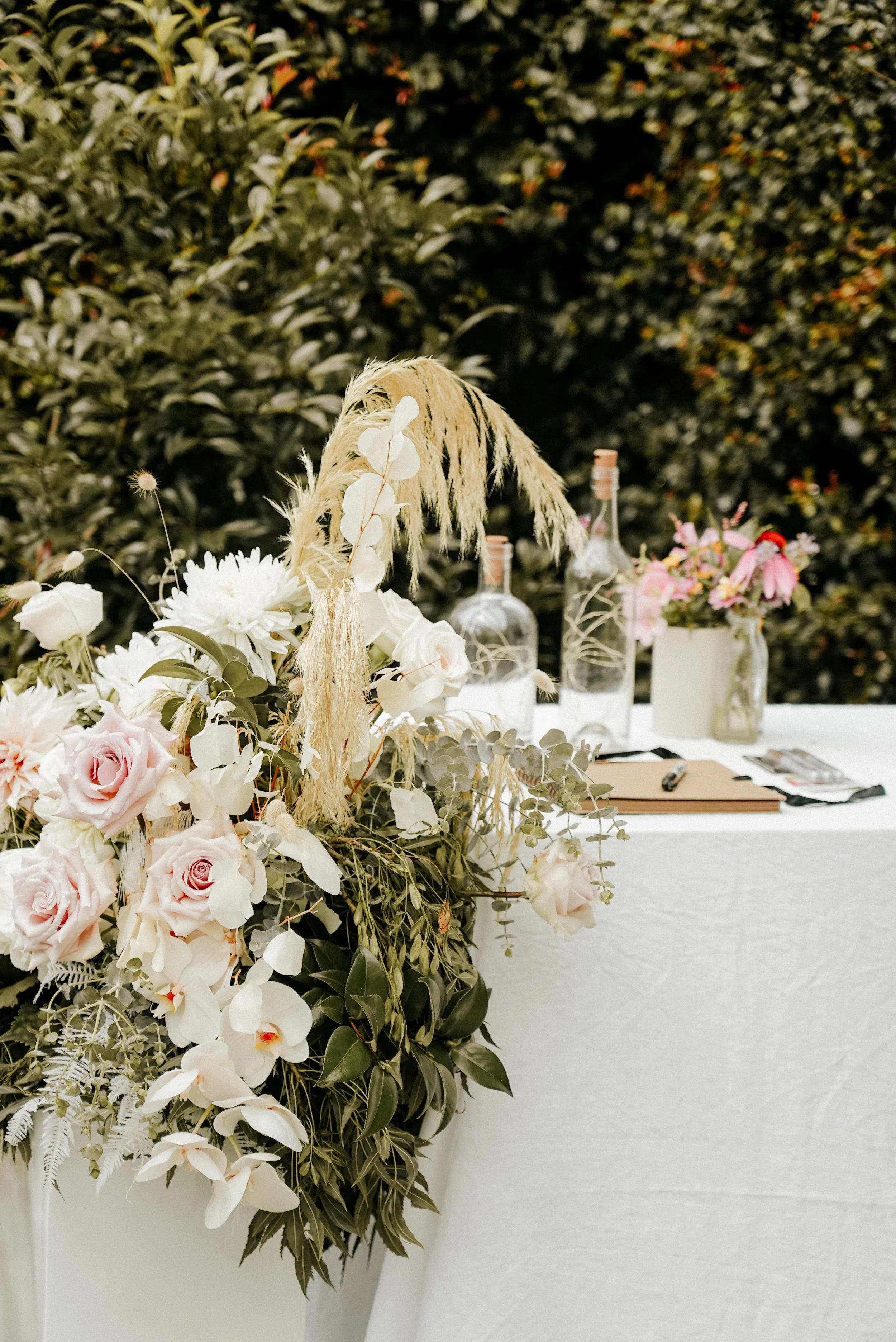Decorative floral arrangement on a white table with bottles and notebooks, outdoors with green foliage in the background.