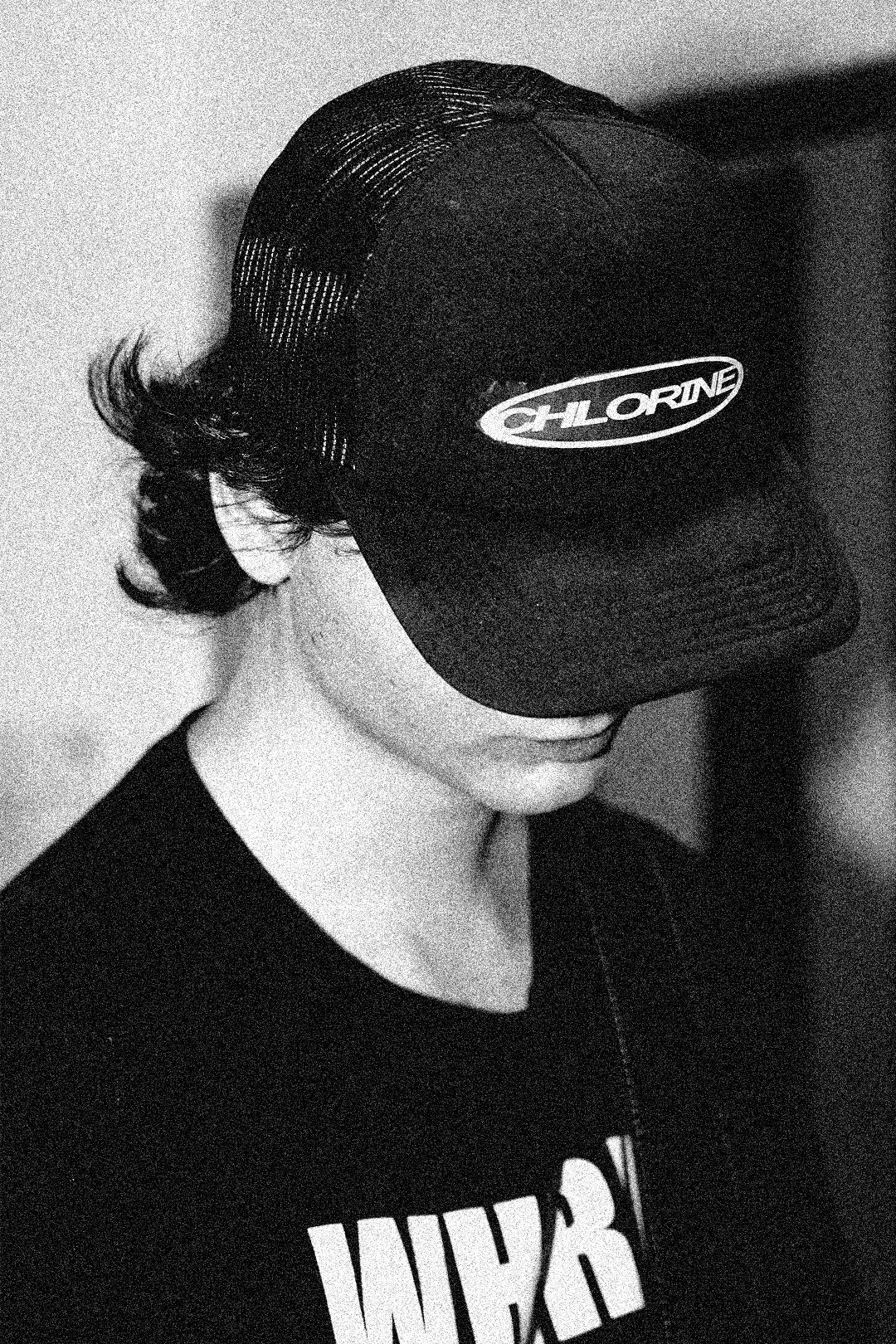 A person wearing a black trucker hat with the logo "Chlorine" and a black T-shirt with partially visible white text, standing with their head slightly down and facing to the side.