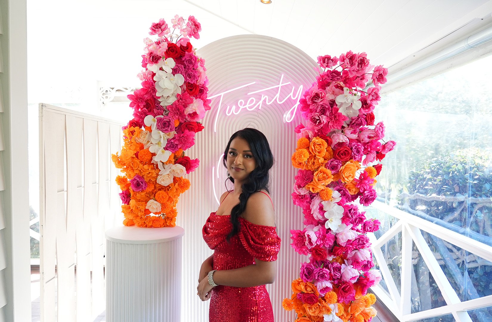 A woman in a red sequined dress standing in front of a floral arch with pink, orange, and white flowers, and a neon sign that says "Twenty".