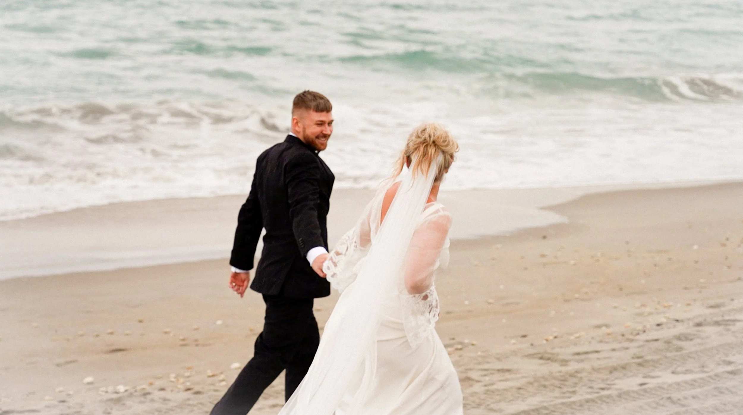 A bride and groom walking hand in hand along the beach, with the ocean waves in the background.