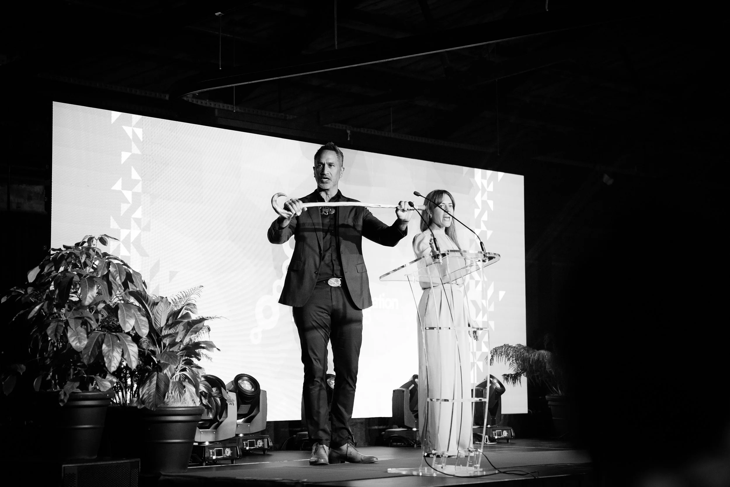 A man and a woman standing at a podium on stage, giving a presentation with a large screen behind them. The man is holding a long object, and the woman is smiling. There are potted plants on the stage.