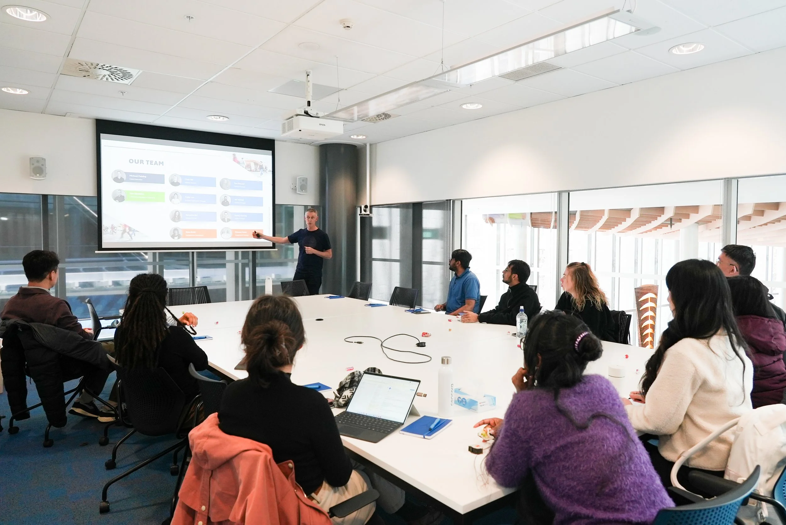 A man gives a presentation to a group of diverse people seated around a large conference table in a modern, well-lit room with large windows and a projector screen displaying a team slide.