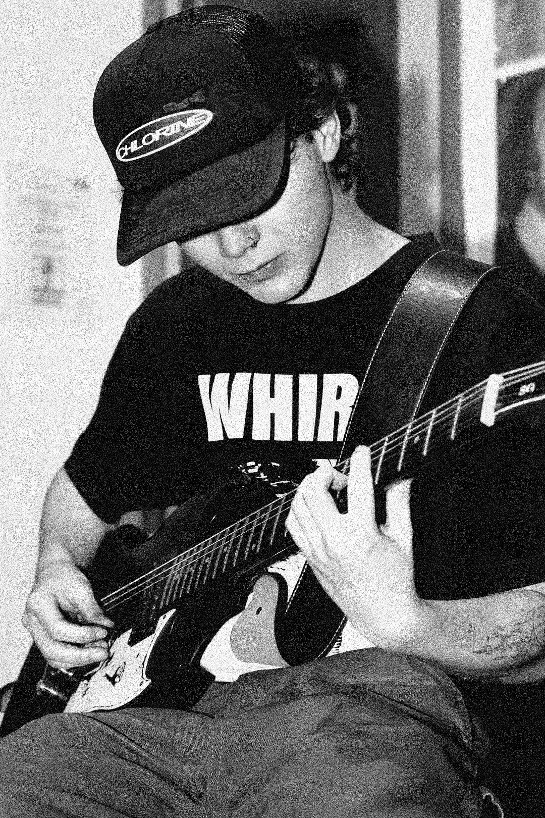 A young person with curly hair wearing a baseball cap, a black t-shirt with the word 'WHIR' visible, and earrings, playing an electric guitar.