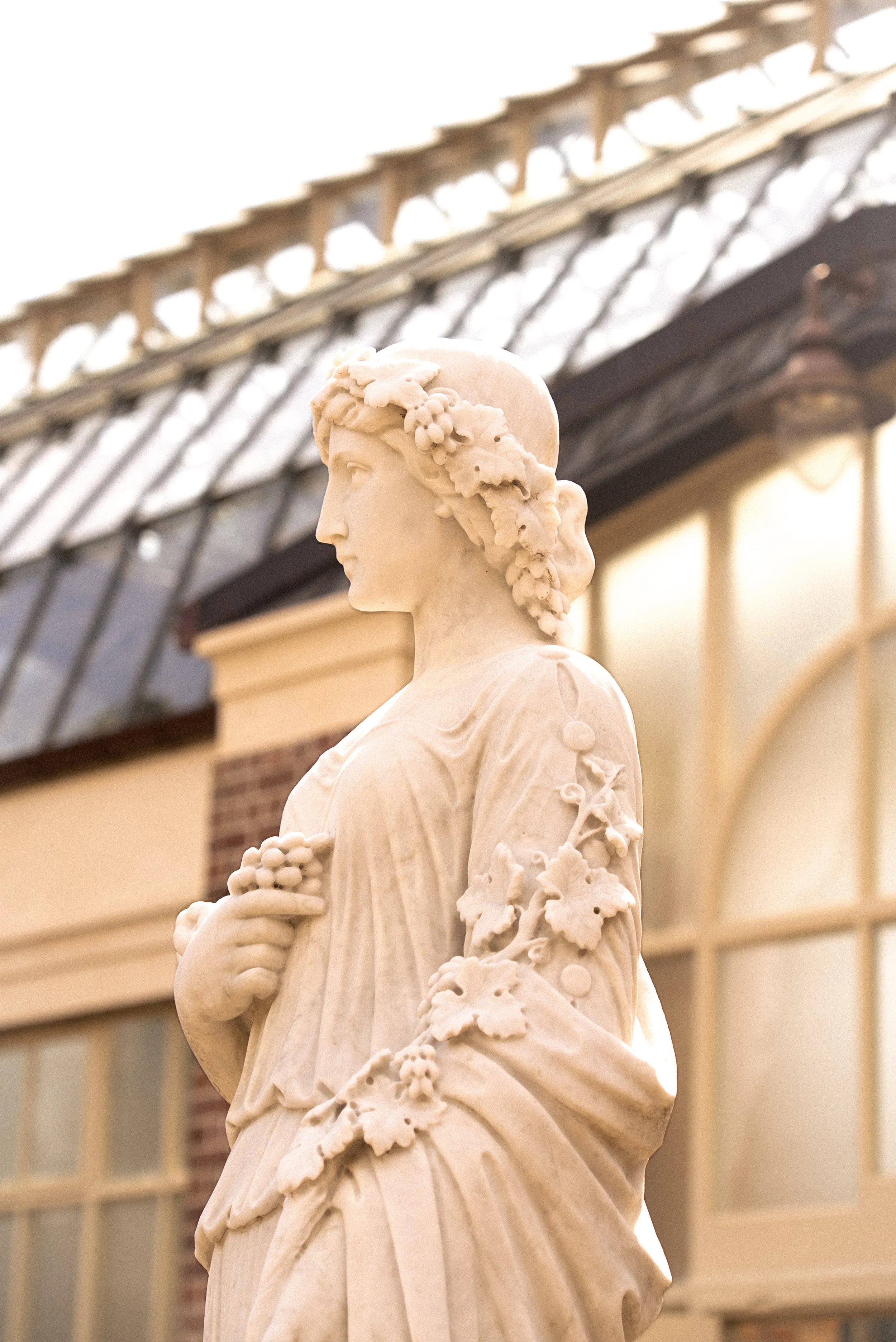 Marble statue of a woman with floral adornments on her head and sleeve, holding a bunch of grapes, against a building with large windows and a glass roof.