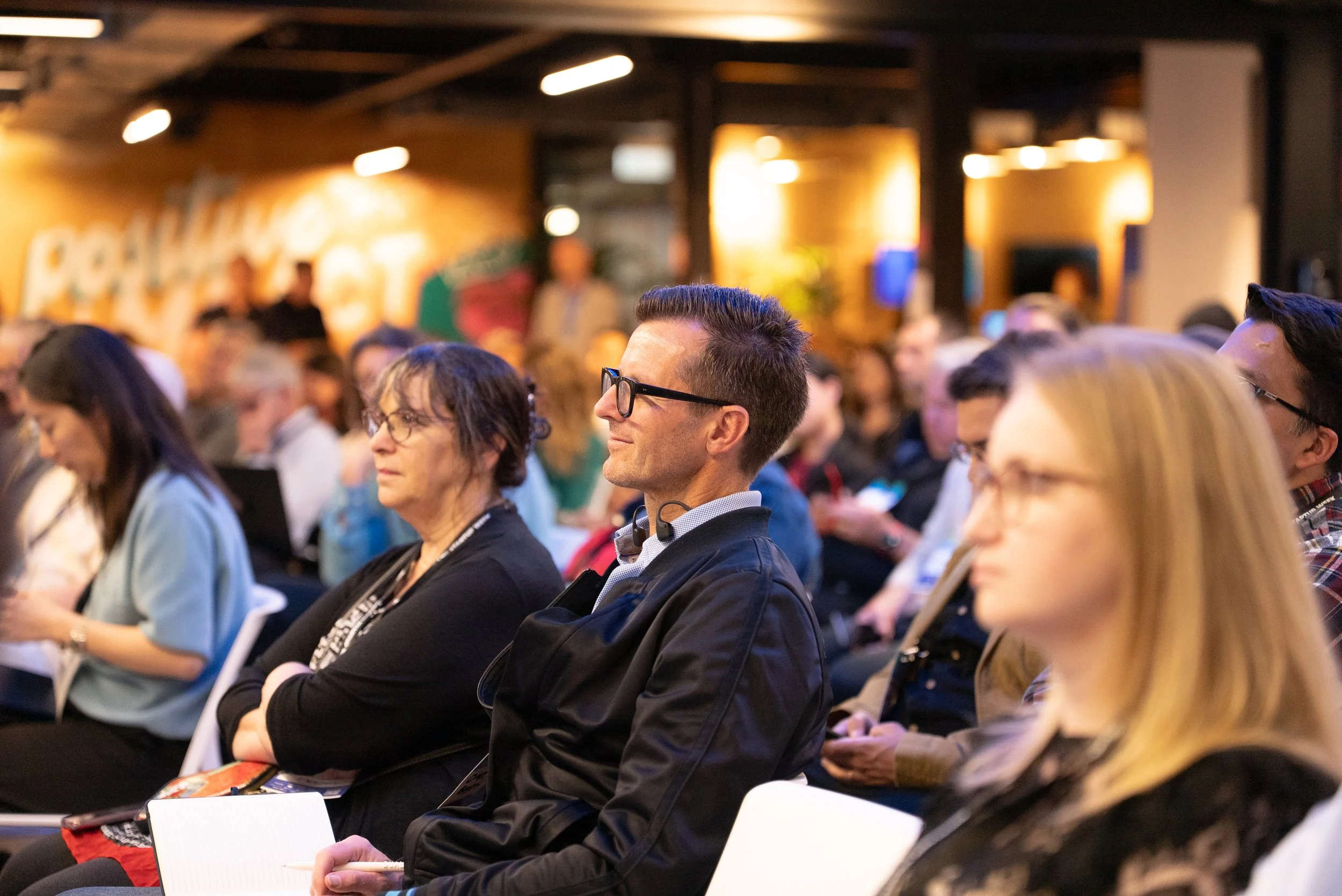 People attending a conference or seminar, sitting and listening attentively, with some taking notes.