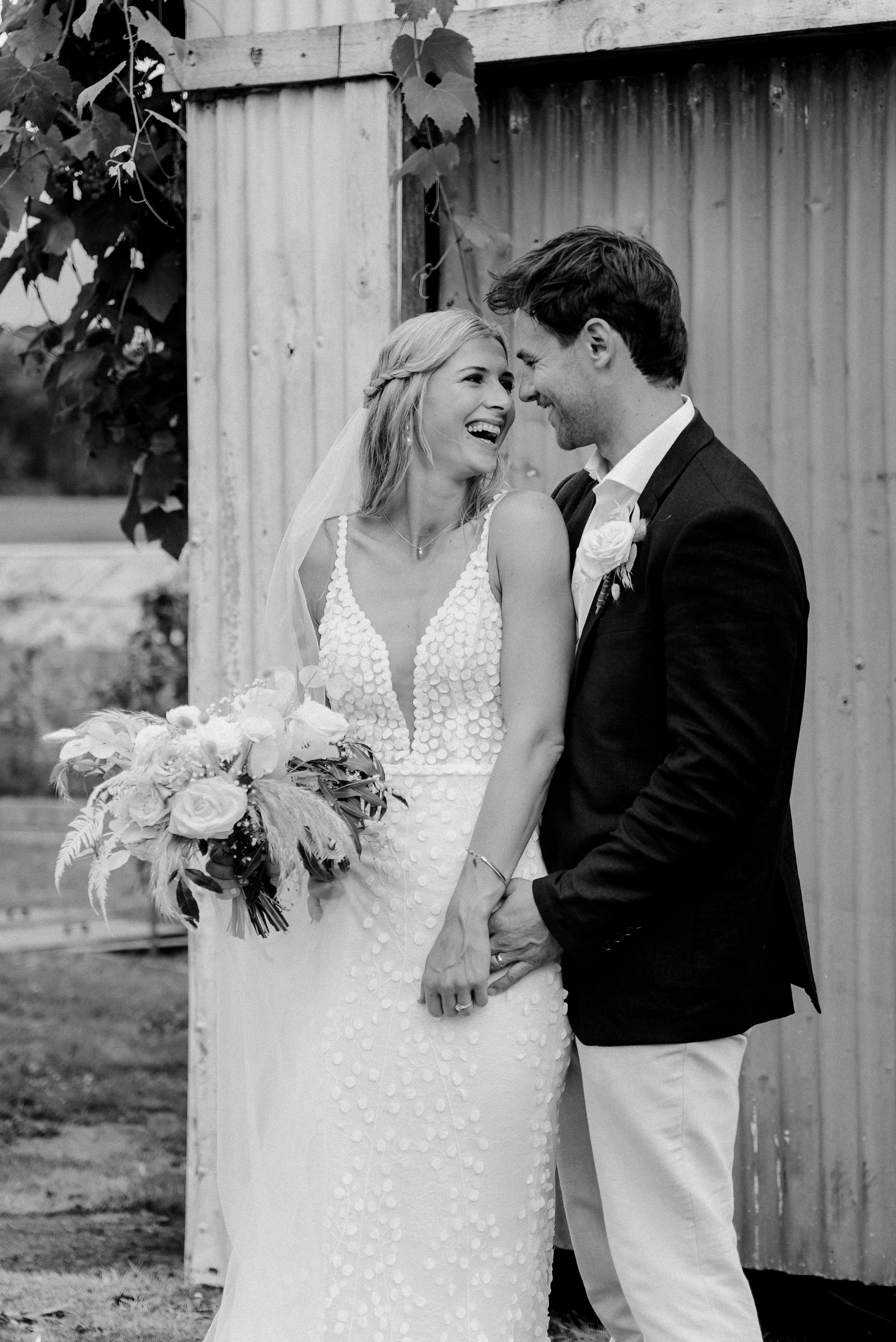 A bride and groom smiling and touching foreheads during their wedding celebration. The bride is holding a bouquet of flowers, and they are standing outdoors in front of a rustic wooden backdrop.