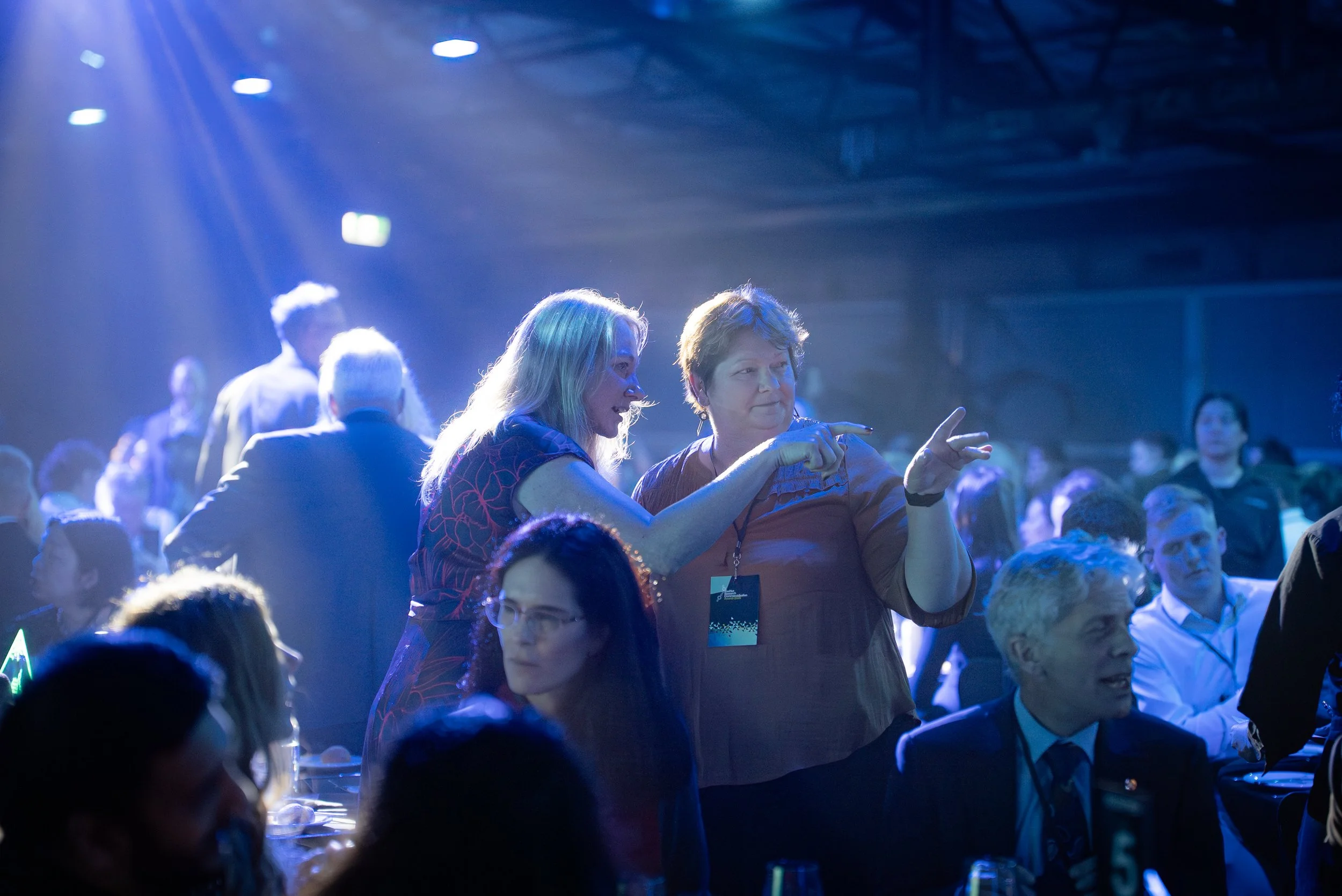 Two women standing and discussing at a crowded event, with people seated at tables and engaging in conversations, illuminated by blue stage lighting.