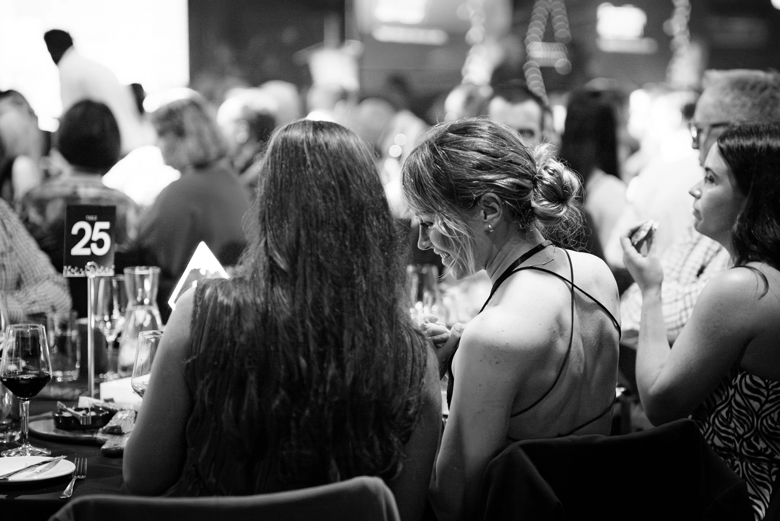 A black-and-white photo of a crowded indoor event with people sitting at tables, some eating and drinking, including wine glasses. The focus is on two women seated at a table, smiling and engaging with each other, with a table number sign '25' visible in the foreground.