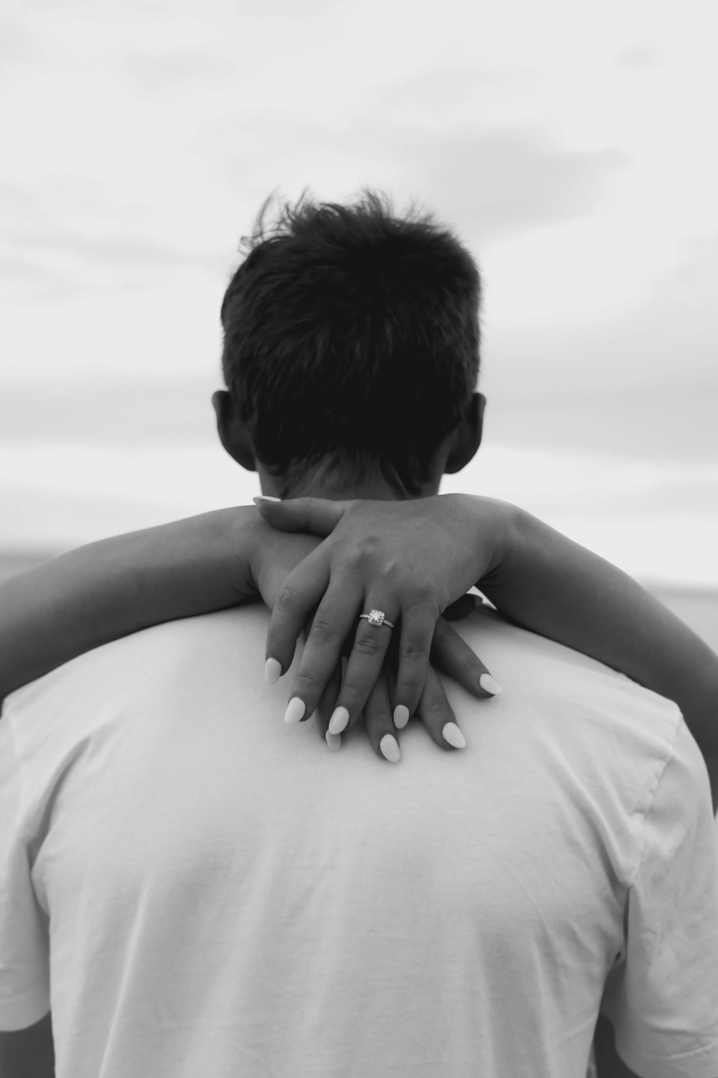 A black and white photo of a woman with her arms wrapped around a man's neck from behind, showcasing her wedding ring.