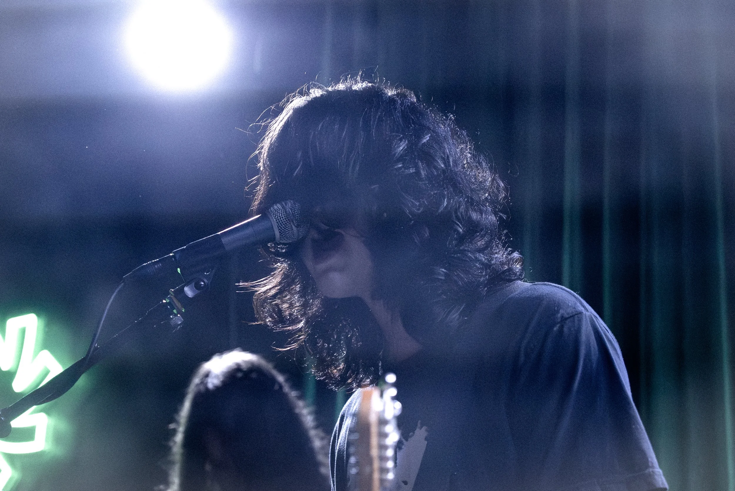 A person performing on stage with curly hair, singing into a microphone, under bright stage lights, with a neon sign in the background.