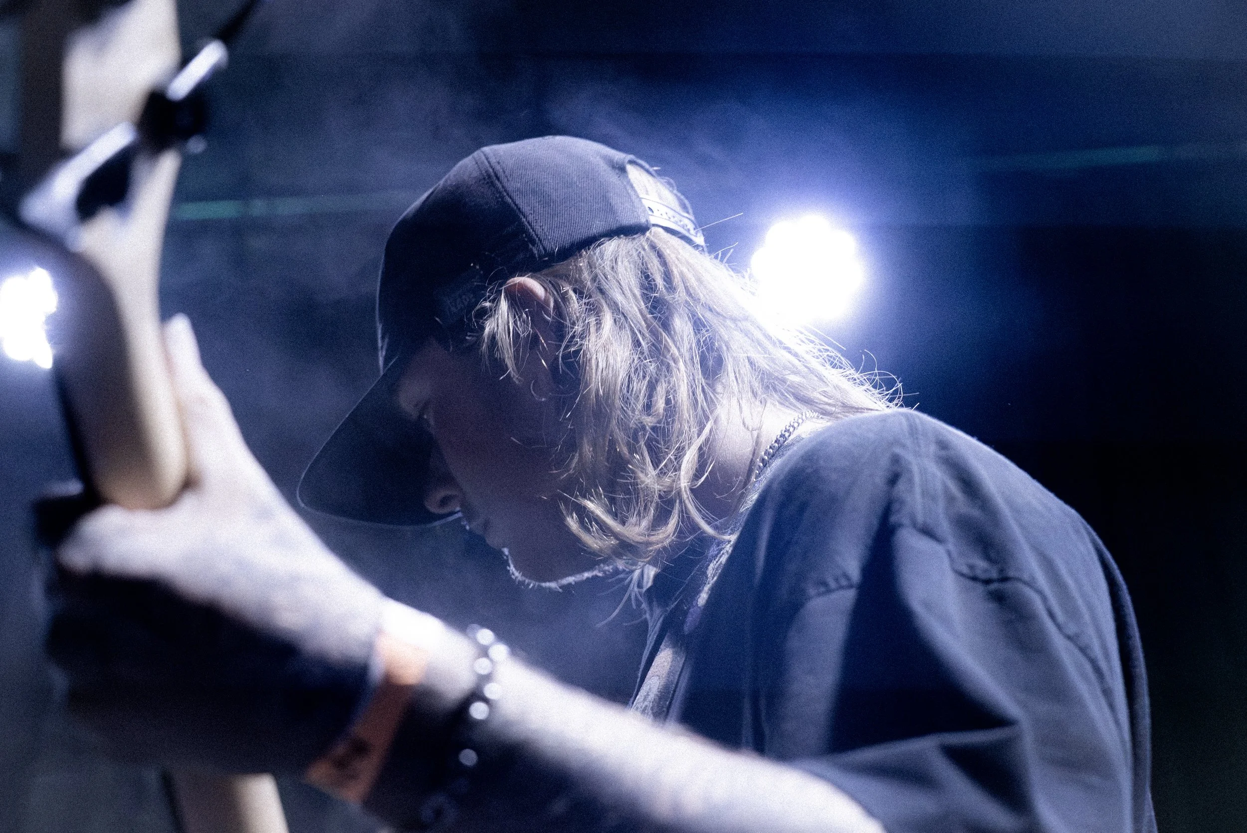 A person with long wavy hair wearing a baseball cap, black T-shirt, and jewelry, leaning on a guitar, illuminated by bright stage lights in a dark setting.