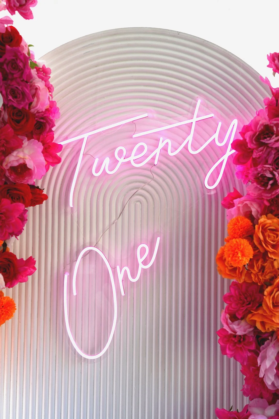 Neon sign displaying the words "Twenty One" in pink, surrounded by pink and orange flowers, against a ribbed white background.