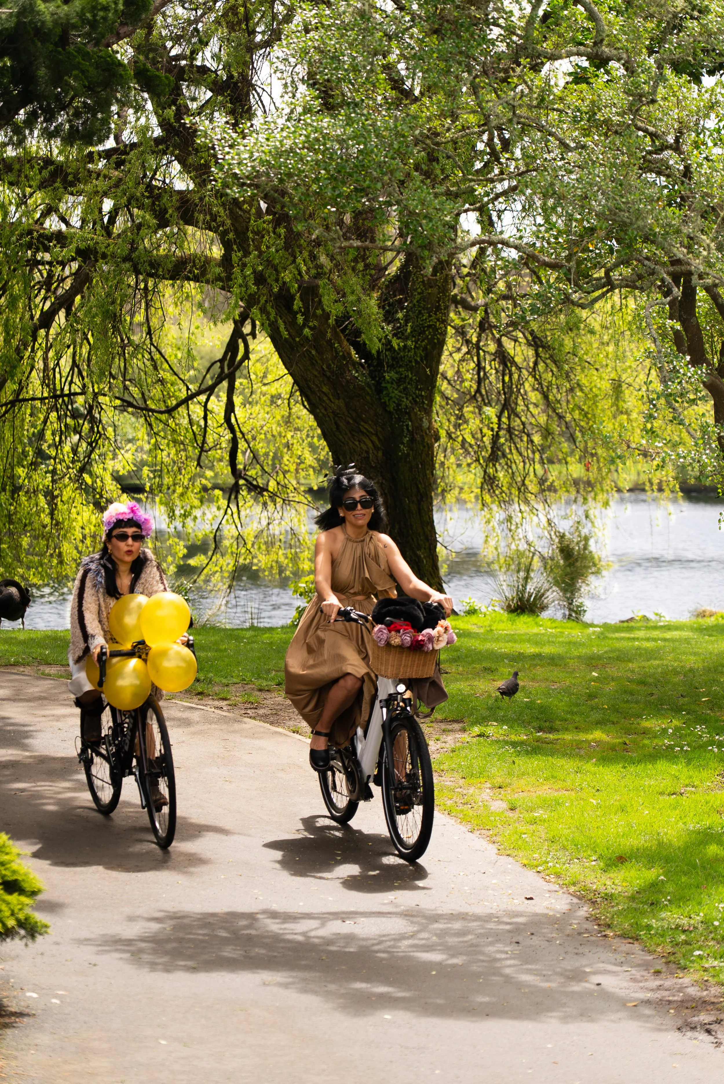 Two women riding bicycles on a path next to a lake, with large trees and grass, one woman wearing a tan dress and sunglasses, the other woman wearing a patterned coat, sunglasses, and pink flowers in her hair, with balloons on her bicycle.