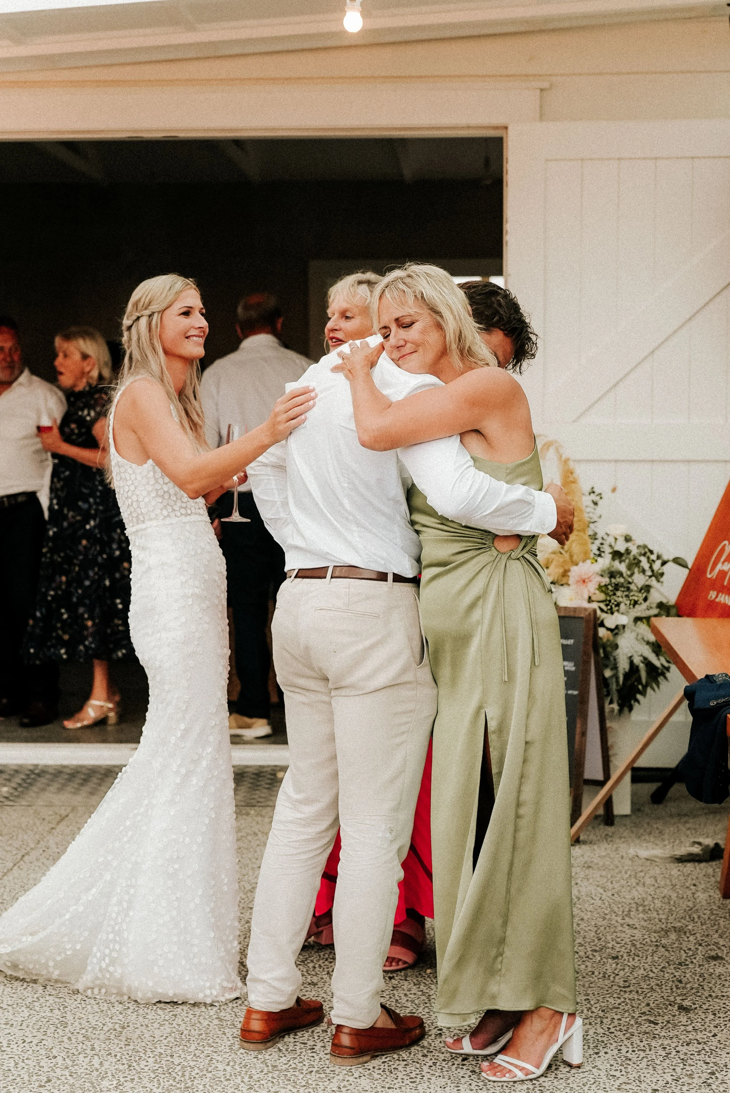 People embracing at a wedding reception, including a woman in a white dress and others with light-colored clothing, in a warmly decorated indoor setting.