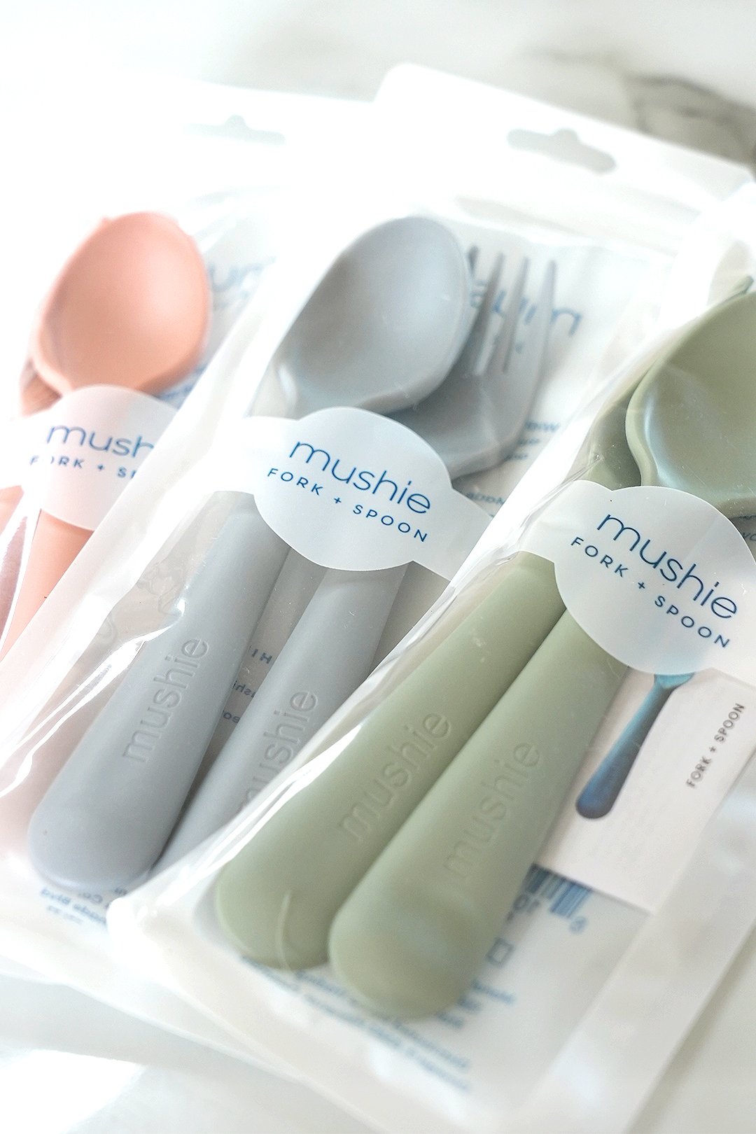 Three sets of silicone utensils with labels, including a pink spatula, light gray fork and spoon, and green fork and spoon, all packaged in clear plastic with 'mushie' branding.