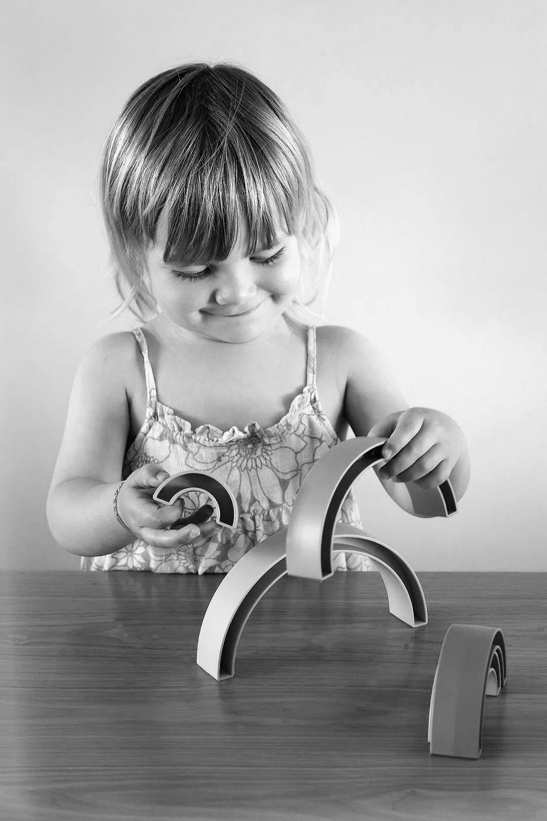 A young girl with short hair, wearing a floral dress, is playing with curved toy blocks on a wooden table. She is smiling gently and looking down at her toys.