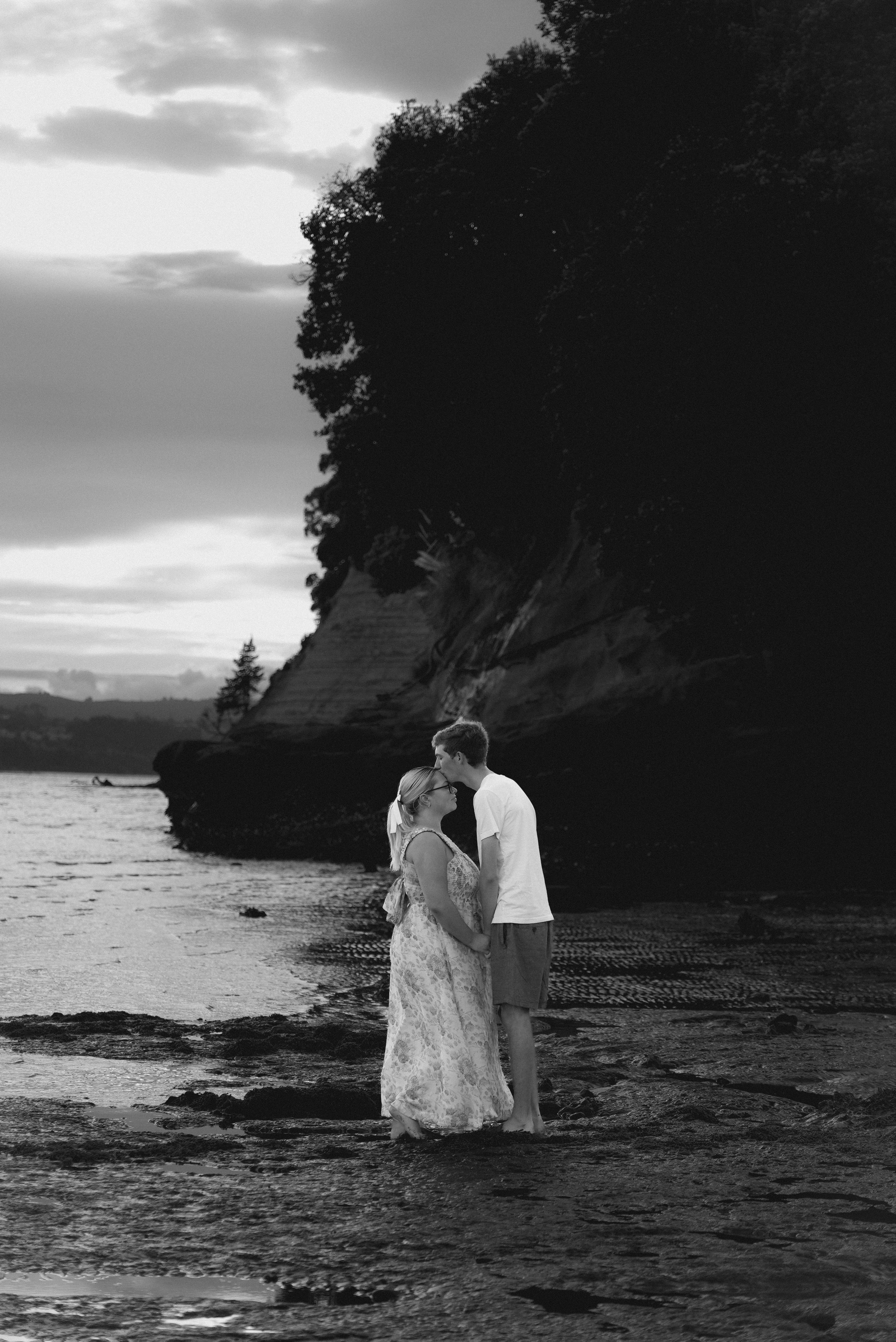 A couple standing in shallow water on the beach, facing each other with foreheads touching, during sunset with cliffs and trees in the background, in black and white.