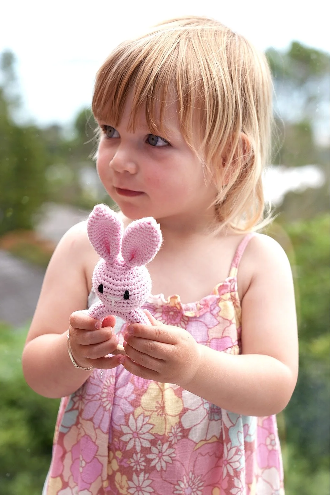 A young girl with red hair, wearing a pink dress with floral patterns, holding a pink crocheted bunny toy with large ears.
