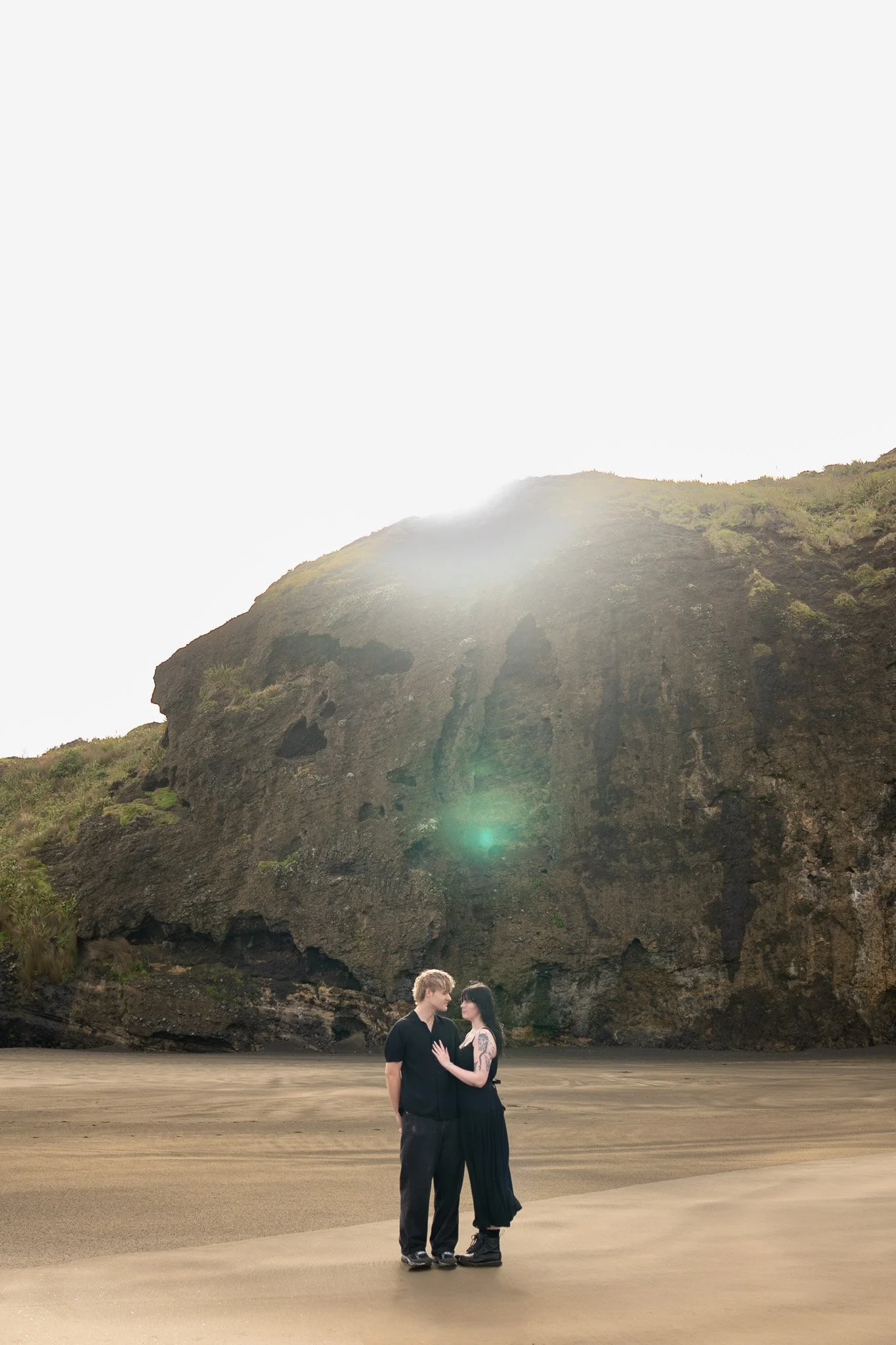 A couple standing close together on a sandy beach with a large cliff in the background and the sun shining brightly overhead.
