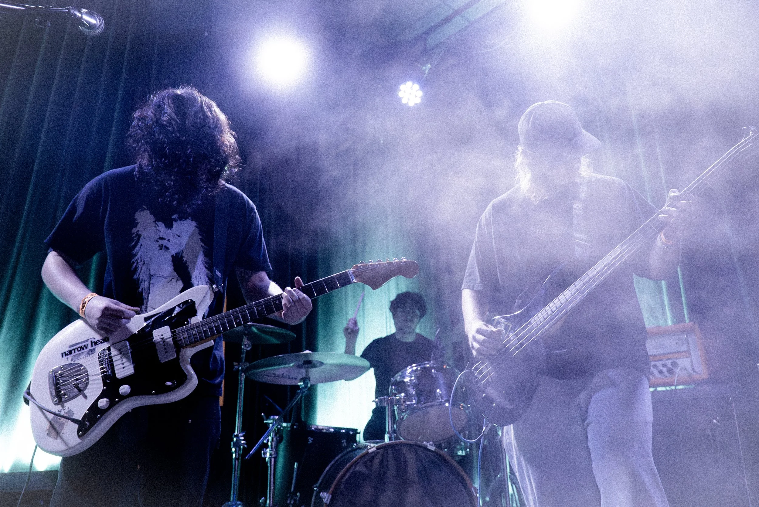 Three musicians performing on stage with guitars and drums, under stage lighting with a green curtain backdrop.